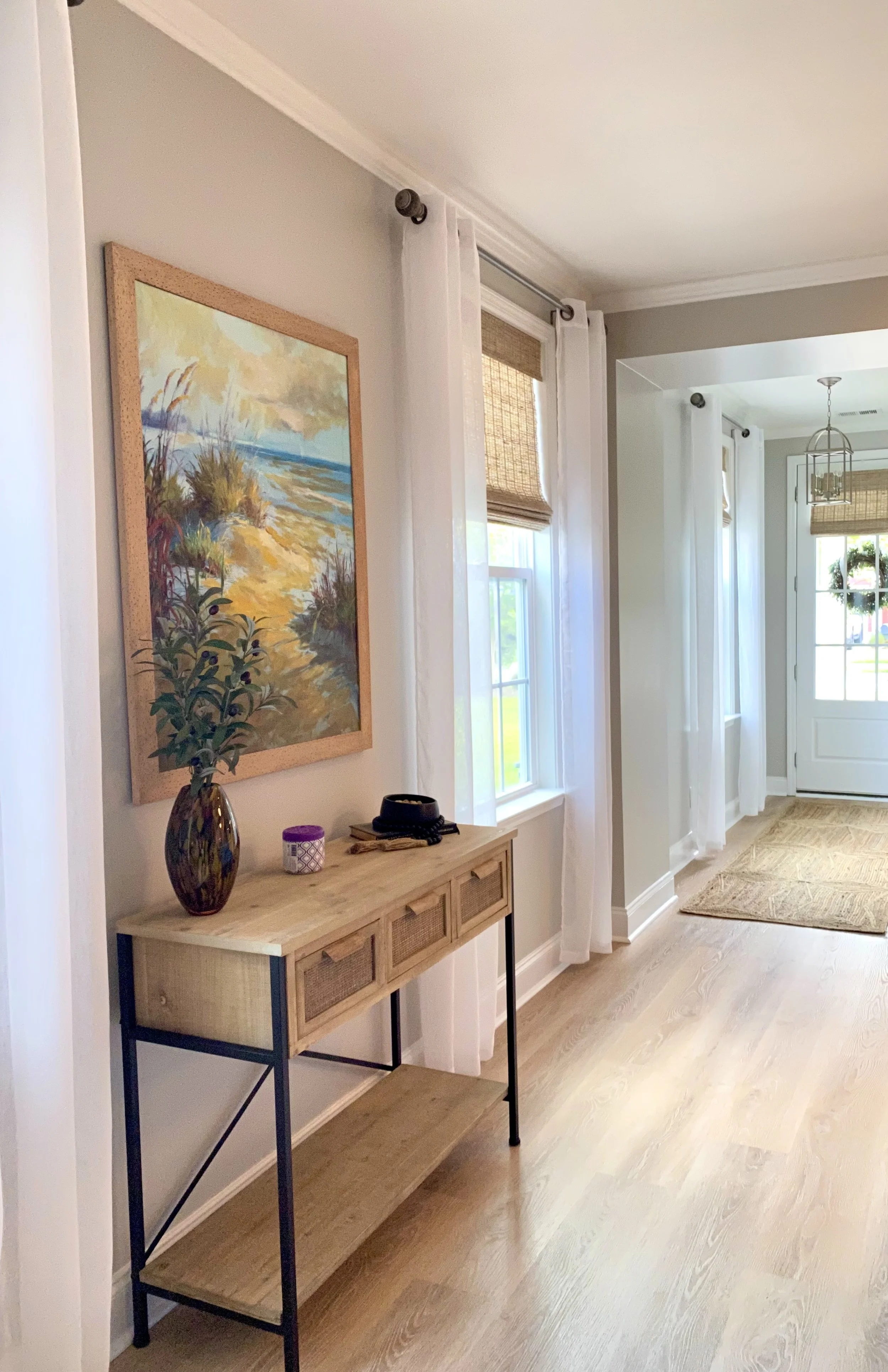 Interior view of a bright, modern living space with light-colored wood flooring, white curtains, and multiple windows with natural light. A decorative painting hangs on the wall above a wooden console table with a vase, a small candle, and other deco