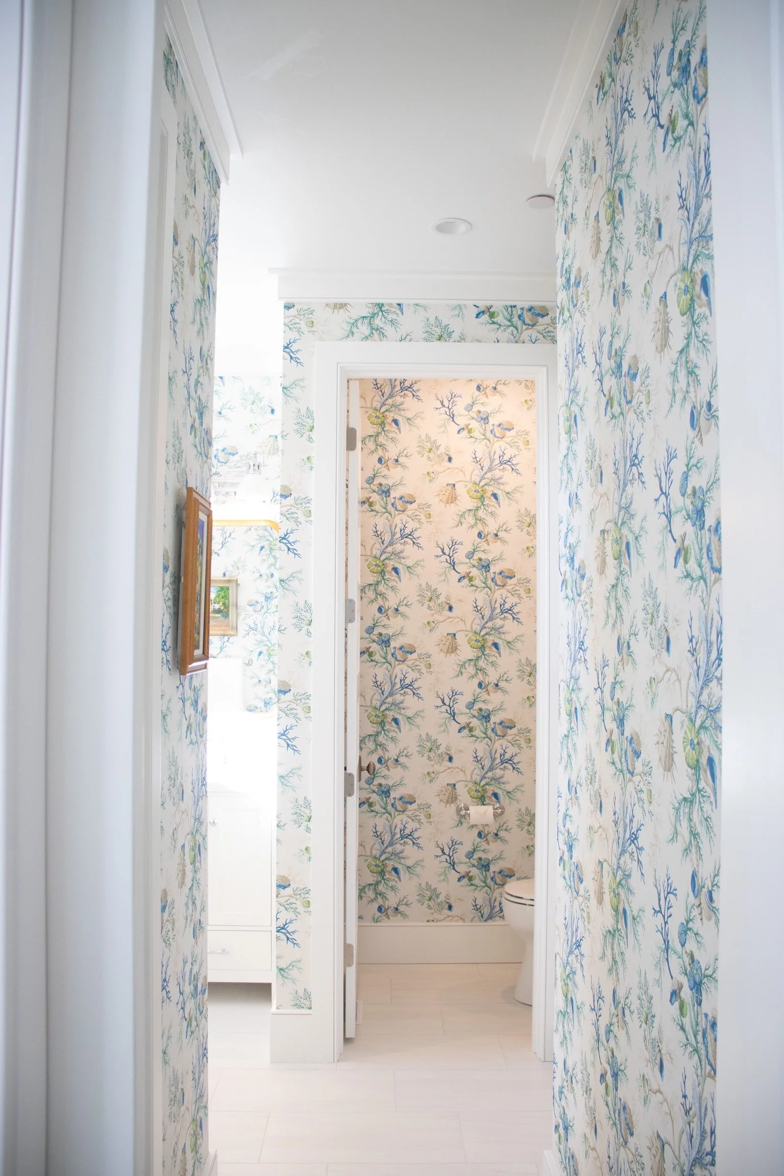 Hallway with blue and green botanical wallpaper leading to a bathroom with similar wallpaper, a white toilet, and a white cabinet with a mirror.