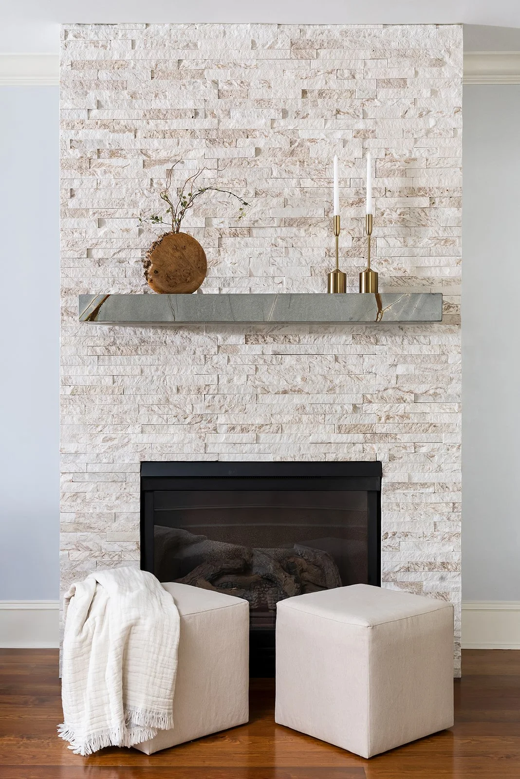 A cozy living room corner with a white stone fireplace, a modern mirrored mantel with a decorative wooden log vase holding a small plant, two tall white candles in gold candleholders, and two beige ottomans with a white throw blanket on one, all on a