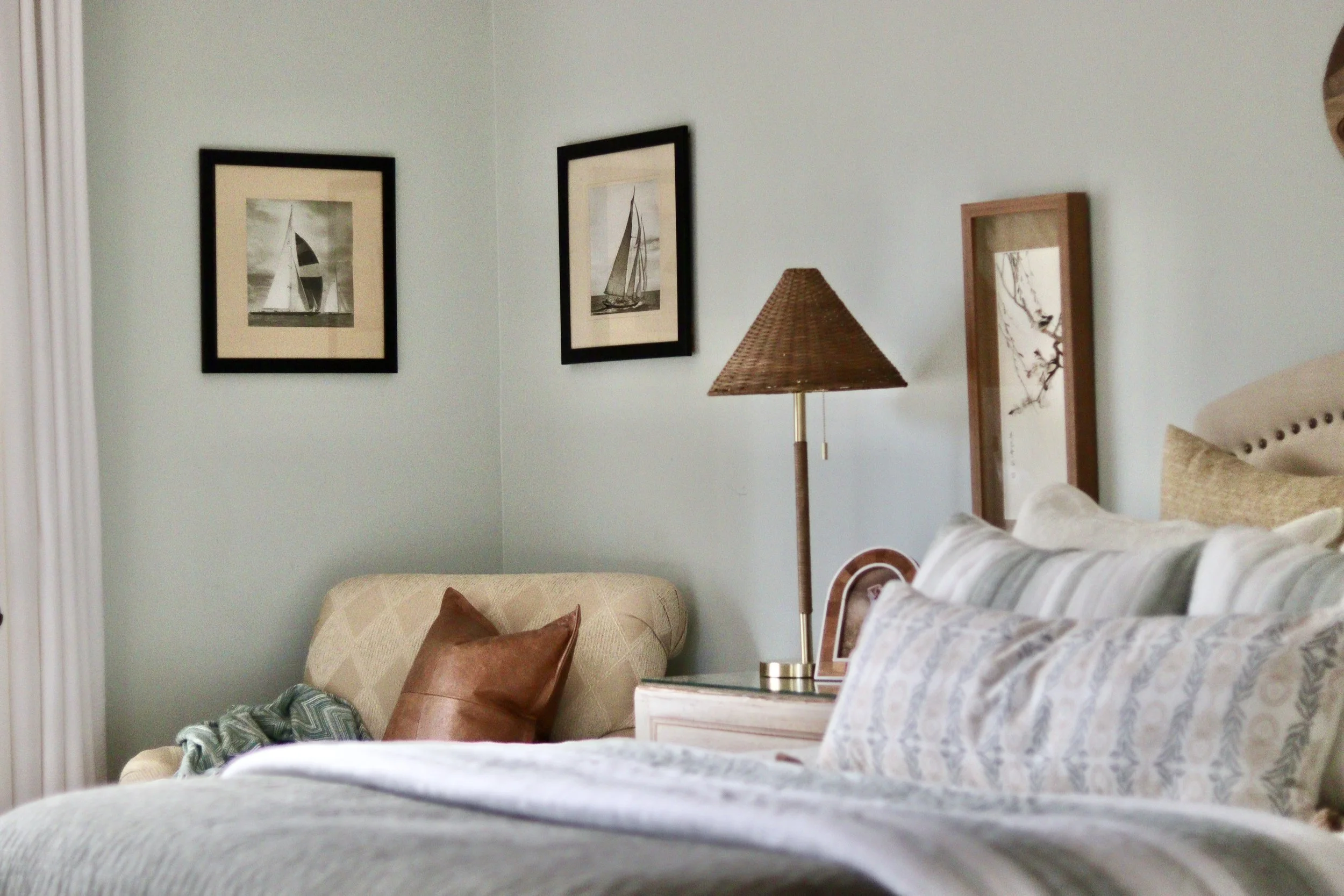 A cozy bedroom with a light green wall, decorated with maritime-themed framed artwork. A beige armchair with brown pillows is in the corner, and a wooden side table holds a wicker lampshade lamp. Part of a bed with a patterned duvet and pillows is vi