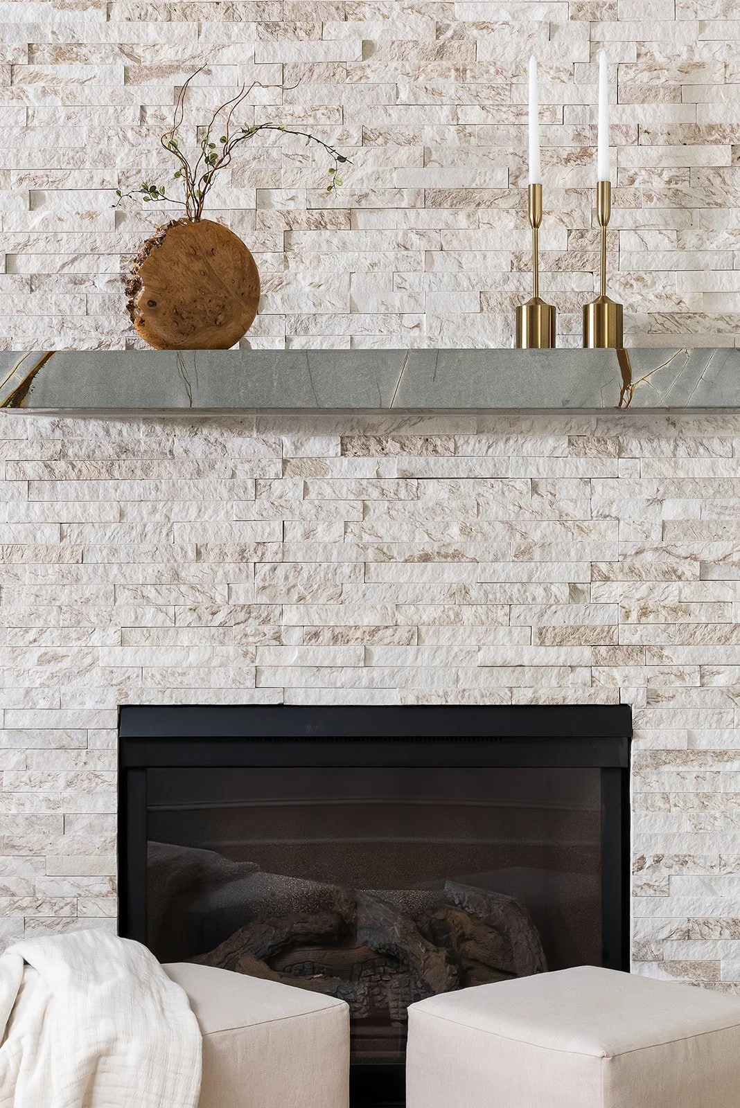 A modern living room fireplace with a light stone wall, a marble mantel, a decorative wood piece with branches and foliage, and two gold candlesticks with white candles.