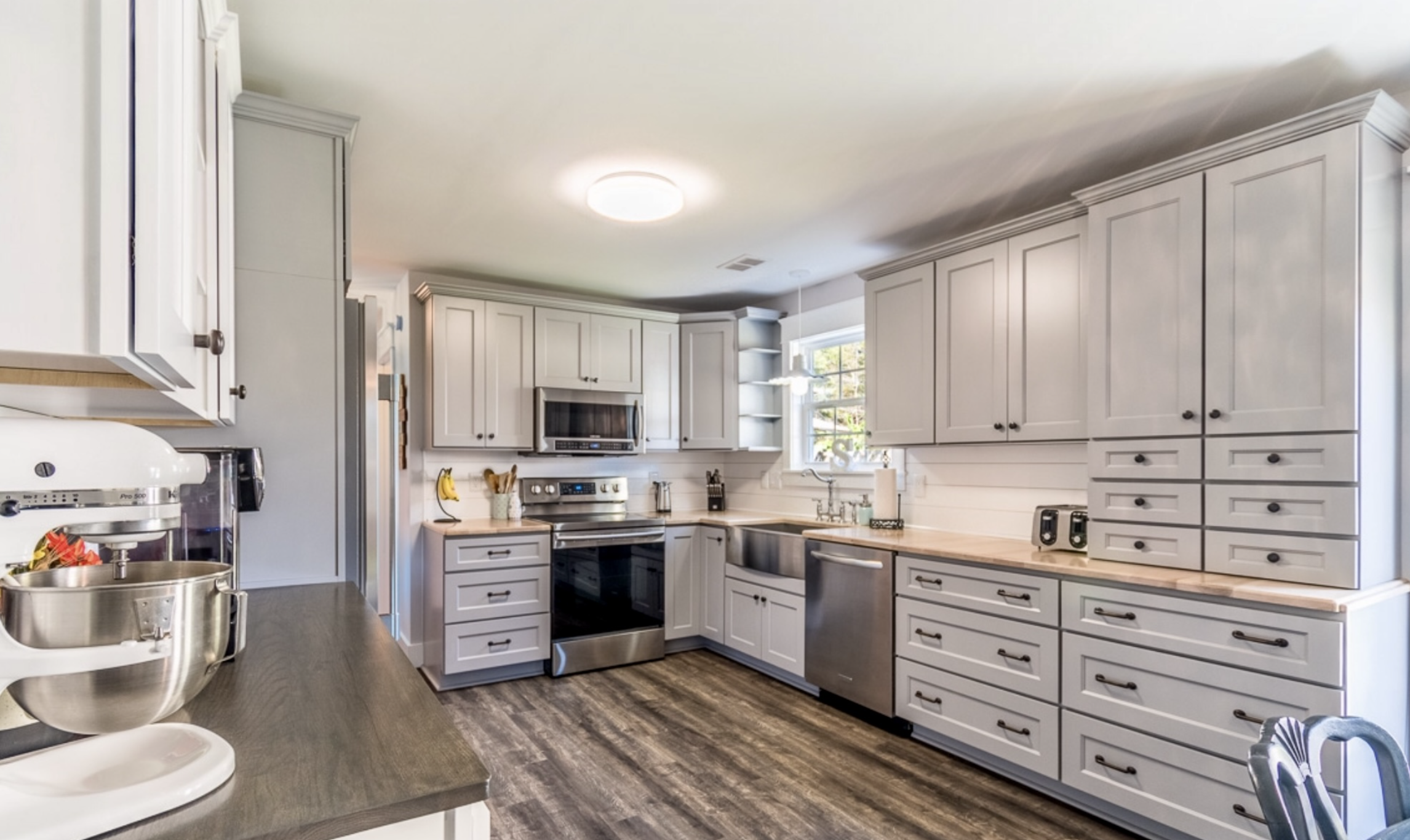 A modern kitchen with white cabinetry, stainless steel appliances, a window above the sink, and a dark wood floor.