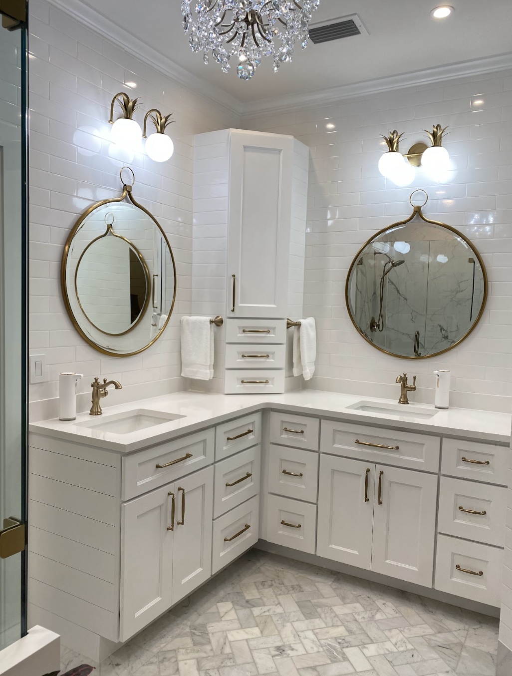 Luxury bathroom with white cabinetry, two round mirrors with gold frames, marble countertops, gold fixtures, two wall-mounted light fixtures, a crystal chandelier, white tiled walls, tiled floor, and towels.