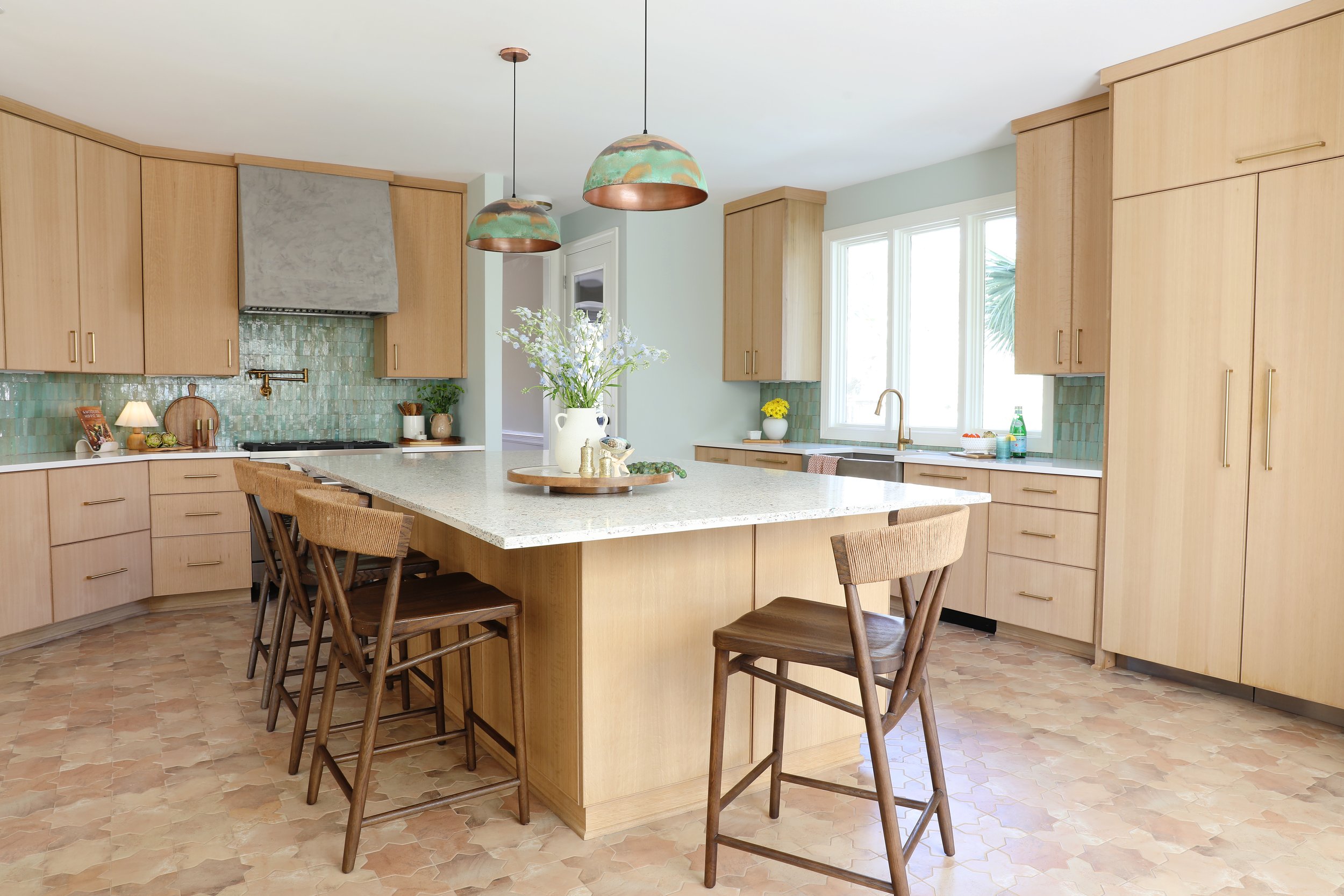 Modern kitchen with light wood cabinetry, a large countertop island with wooden chairs, green tiles behind the stove, and large windows allowing natural light, decorated with a vase of white flowers and small green accents.
