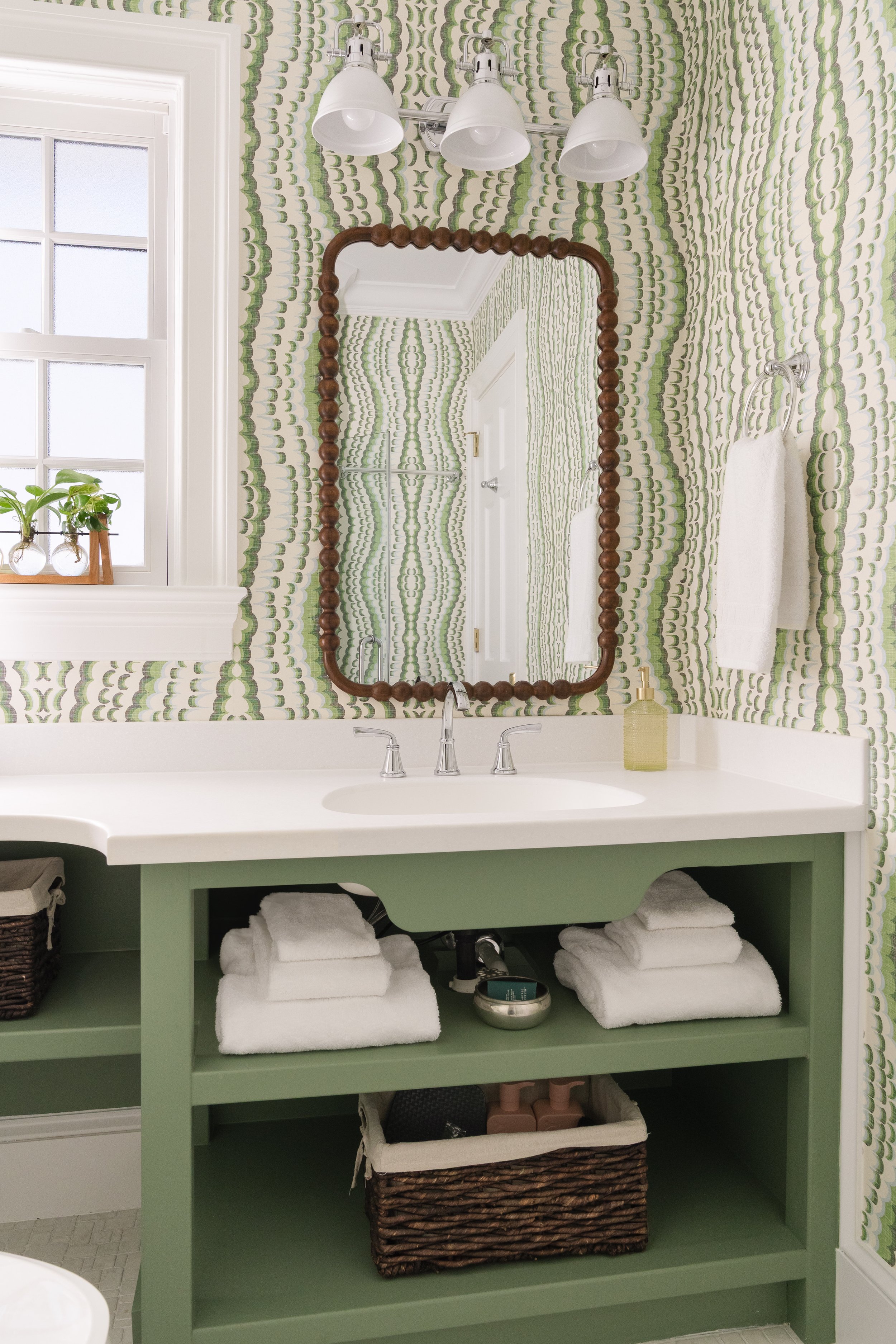 A bathroom vanity with a white countertop, a wide mirror with a wooden bead frame, and a mint green cabinet below storing rolled white towels and a wicker basket. The wall behind has green and white patterned wallpaper, and there is a chrome faucet o