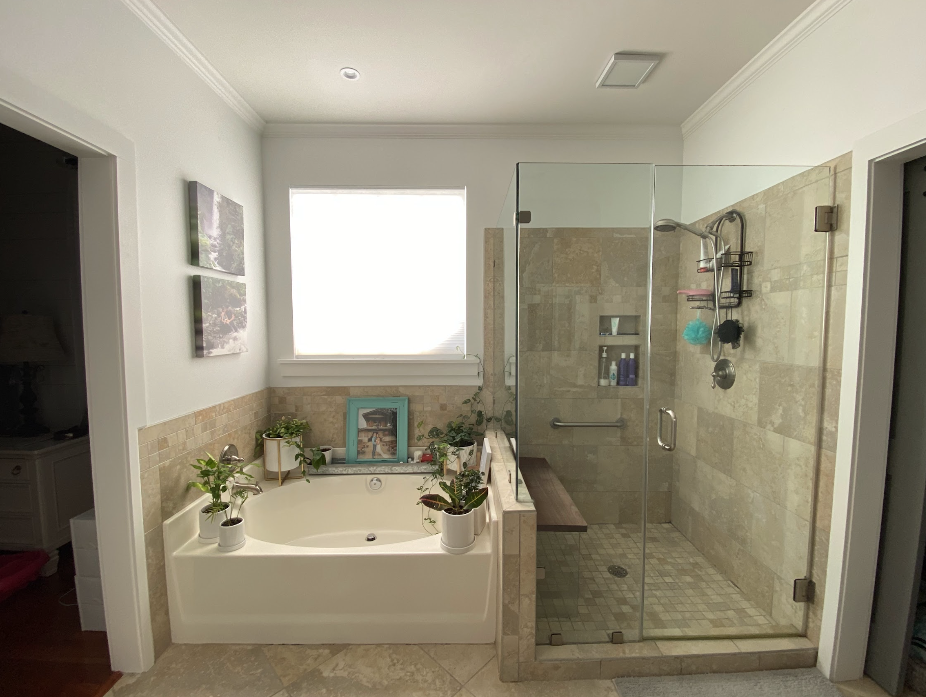 A bathroom with a corner bathtub with plants and pictures around it, adjacent to a walk-in shower with glass doors and beige tiles.