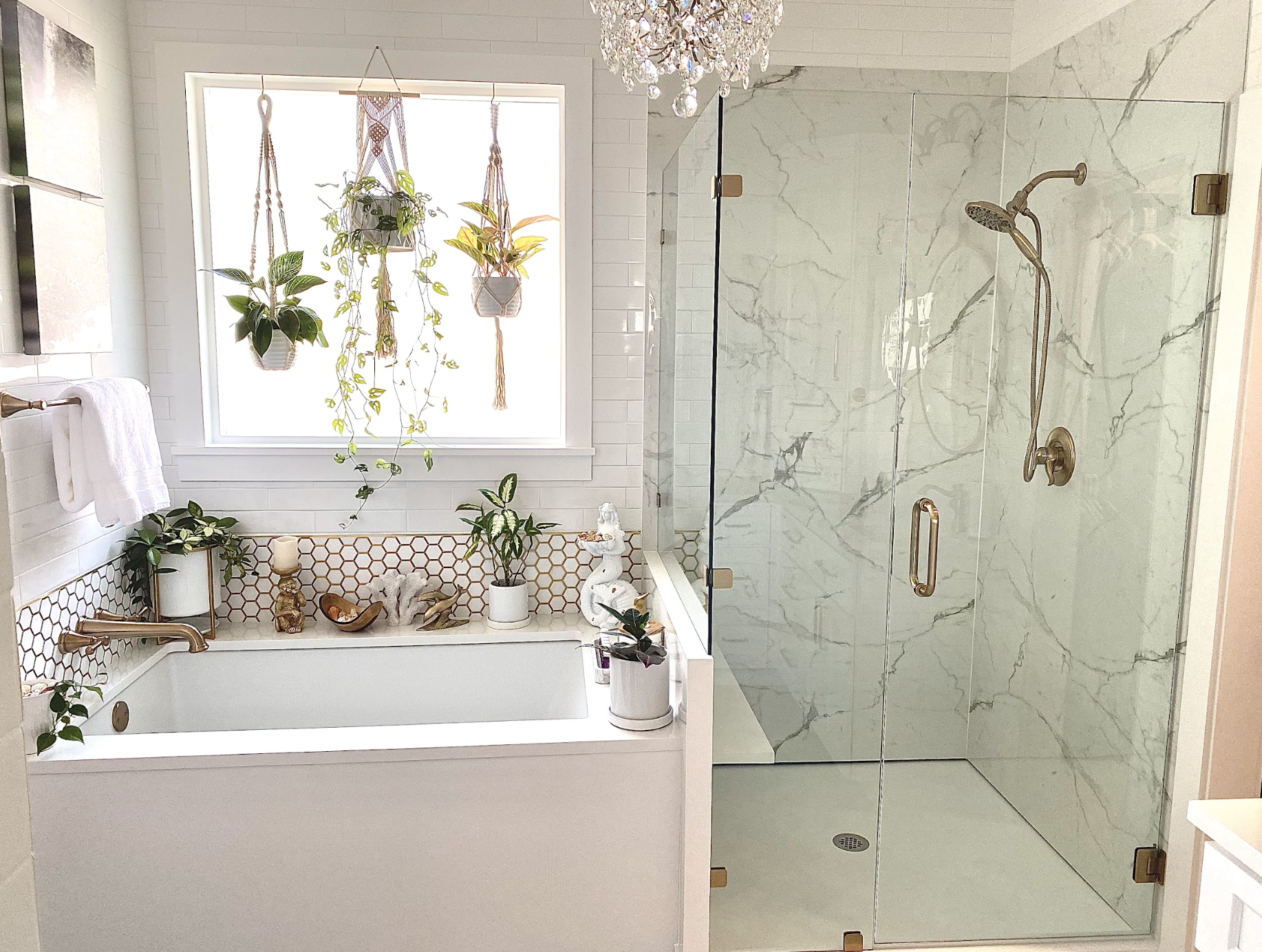 A bathroom with a white bathtub filled with decorative items, plants, and candles, a large window with hanging plants, and a glass-enclosed shower with marble walls.