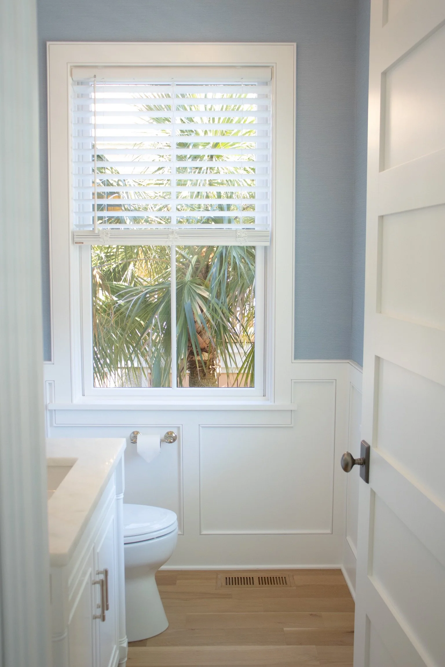 A small bathroom with a white toilet, a white vanity with a faucet, a large window with white blinds showing a palm tree outside, and a wooden floor with a vent. The walls are painted light blue and there is a white door partially open.