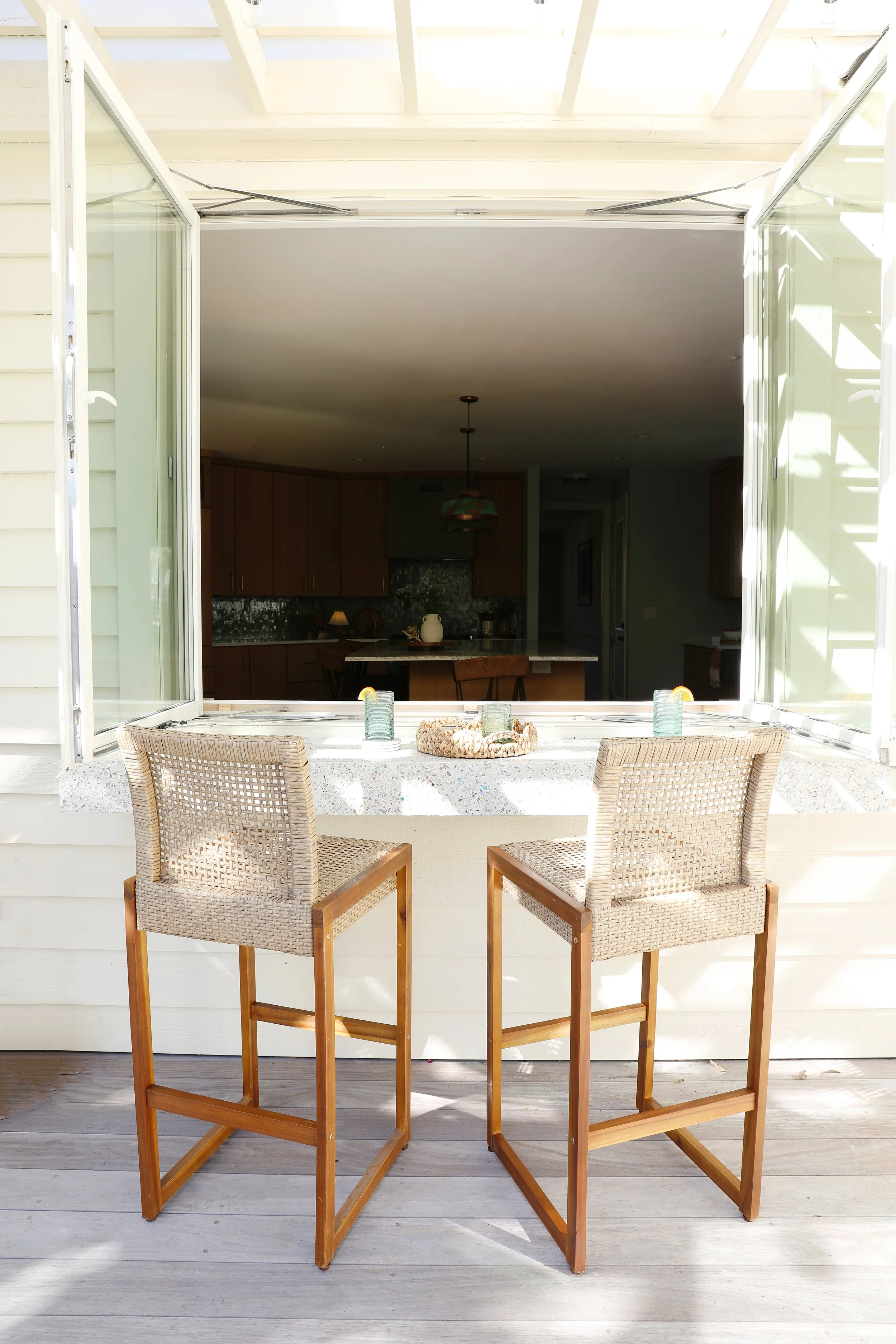 Open window with two wicker chairs and a countertop on a porch, overlooking a kitchen interior.