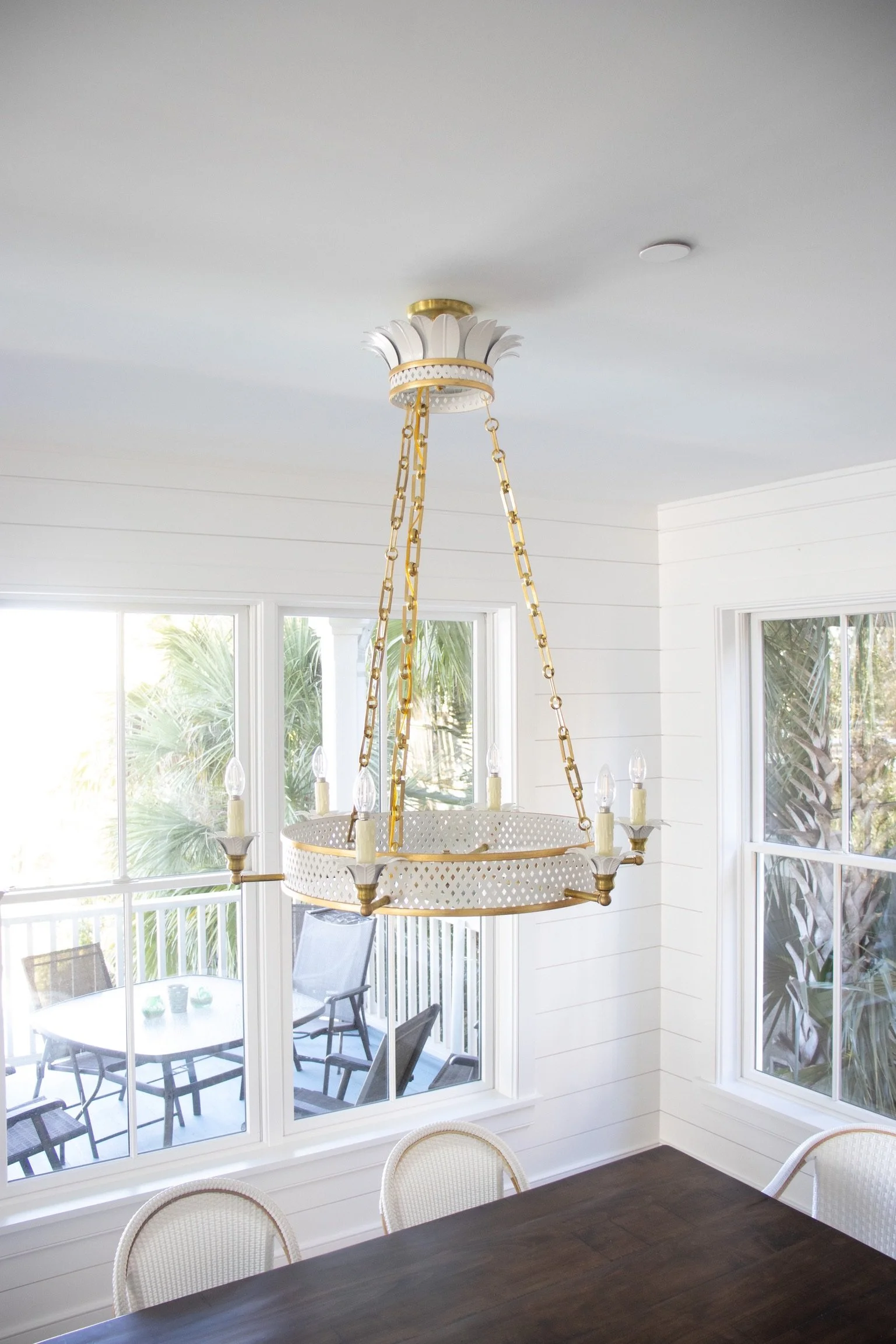 A chandelier hanging from a white ceiling in a room with large windows, white walls, and a dark wooden dining table with chairs.
