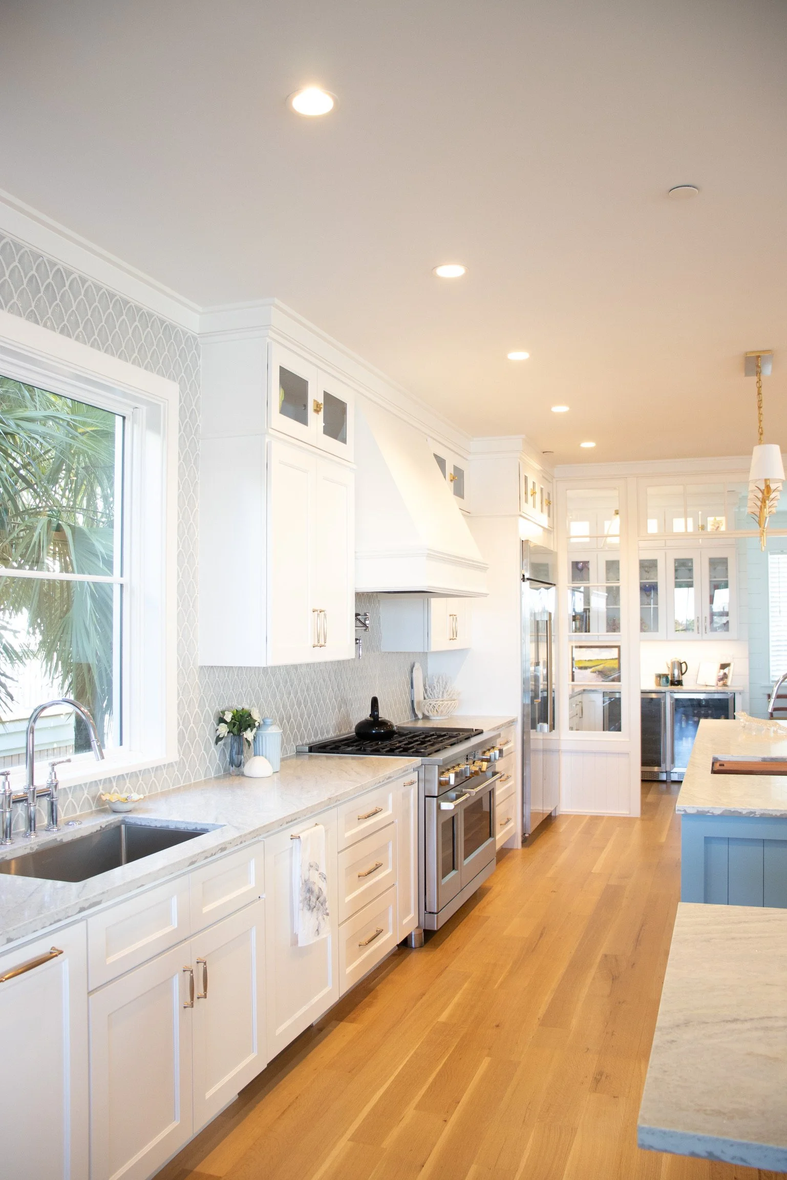 Modern white kitchen with wooden flooring, white cabinets, marble countertops, stainless steel stove, and a large window with a view of greenery.