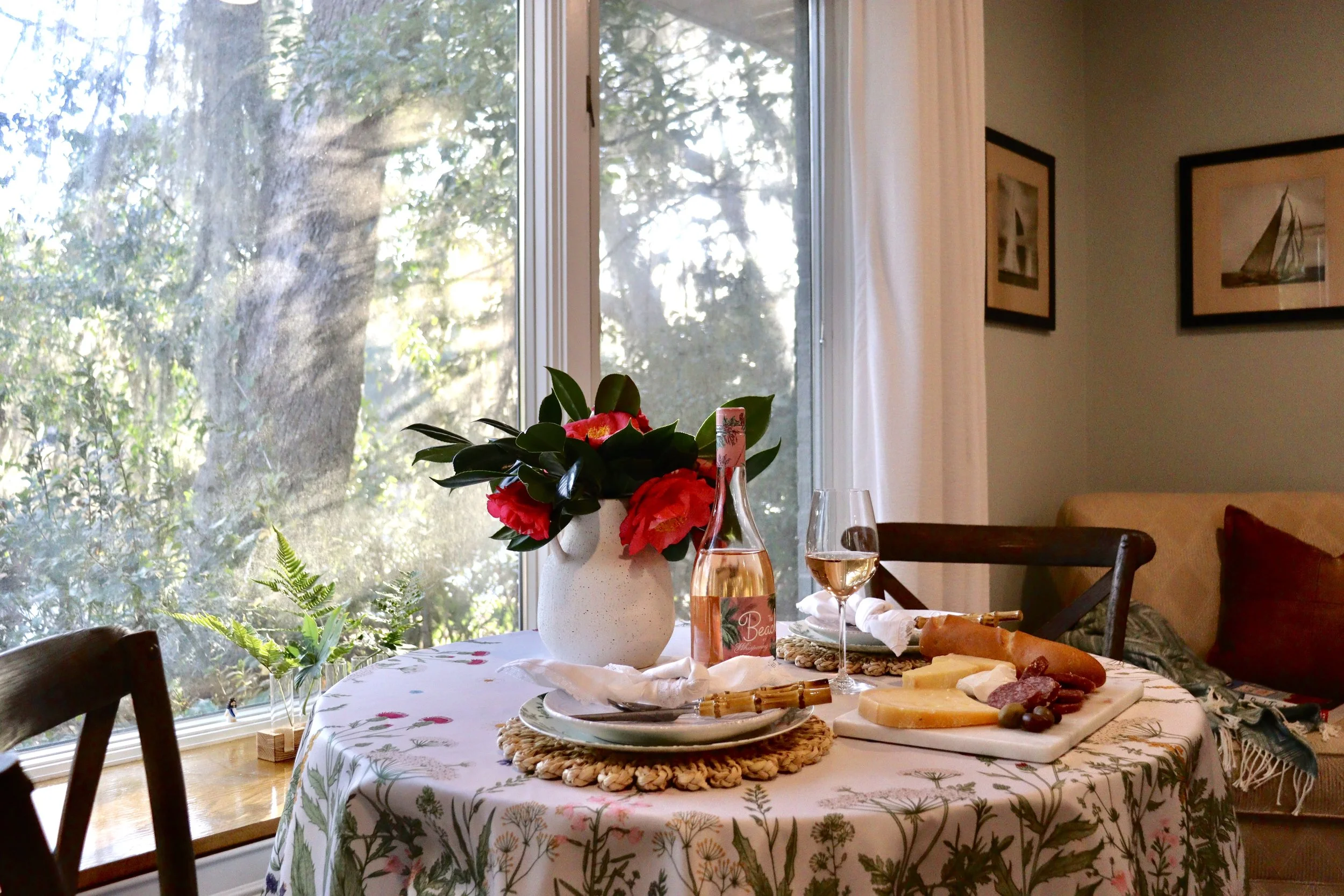 A dining table with a white tablecloth featuring green botanical prints, set with plates, cutlery, and a charcuterie board, with a vase of pink and red flowers, a bottle of rosé wine, and a wine glass. The background shows a large window with a view 