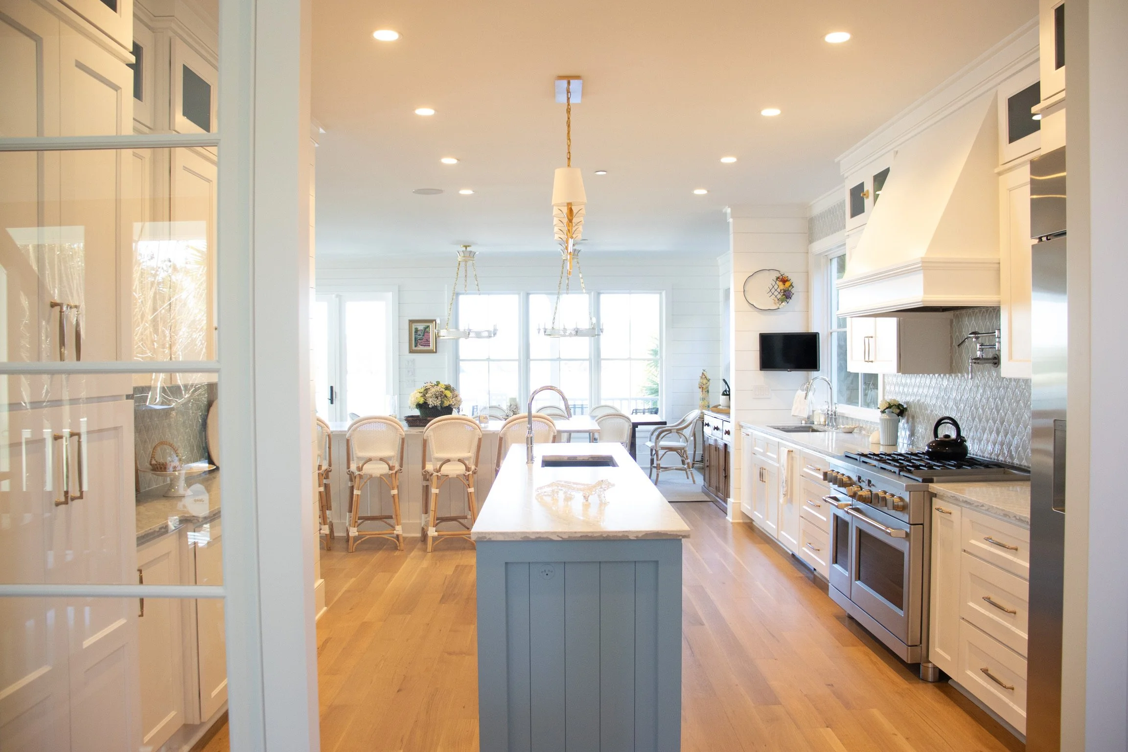 A spacious, bright kitchen with white cabinets, a large central island with a white marble countertop, stainless steel appliances, and wooden flooring. In the background, a dining area with a table and chairs is visible, illuminated by natural light 