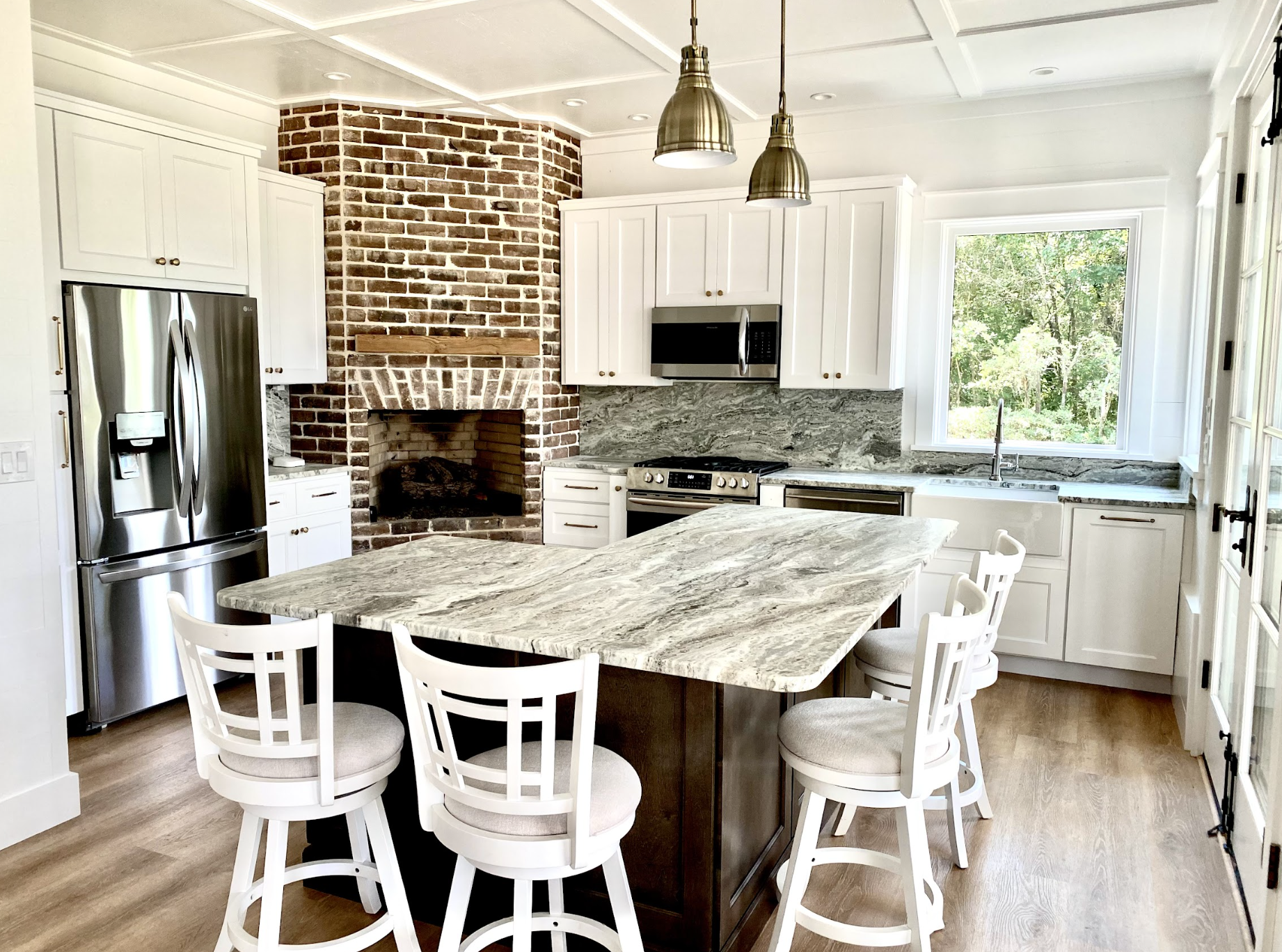 A modern kitchen with white cabinets, a marble island, stainless steel refrigerator, microwave, and stove, brick fireplace, large window with a view of trees, and wooden flooring.