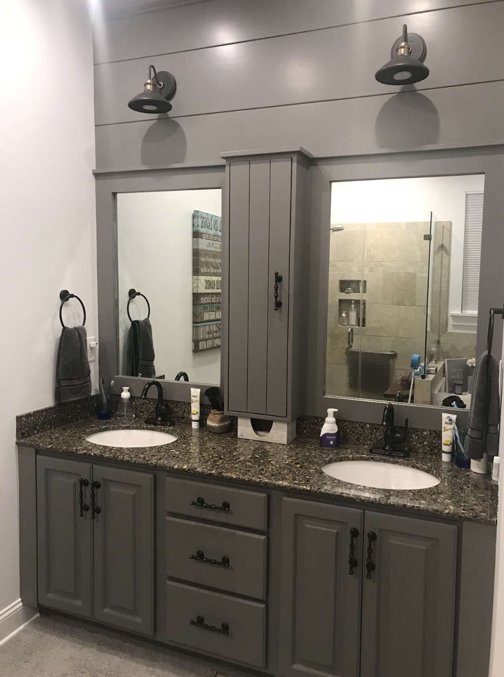 Bathroom with a double vanity, granite countertop, two oval sinks, large mirror with two wall-mounted light fixtures, gray cabinetry, hand towels, and toiletries, with a view of the shower behind the mirror.
