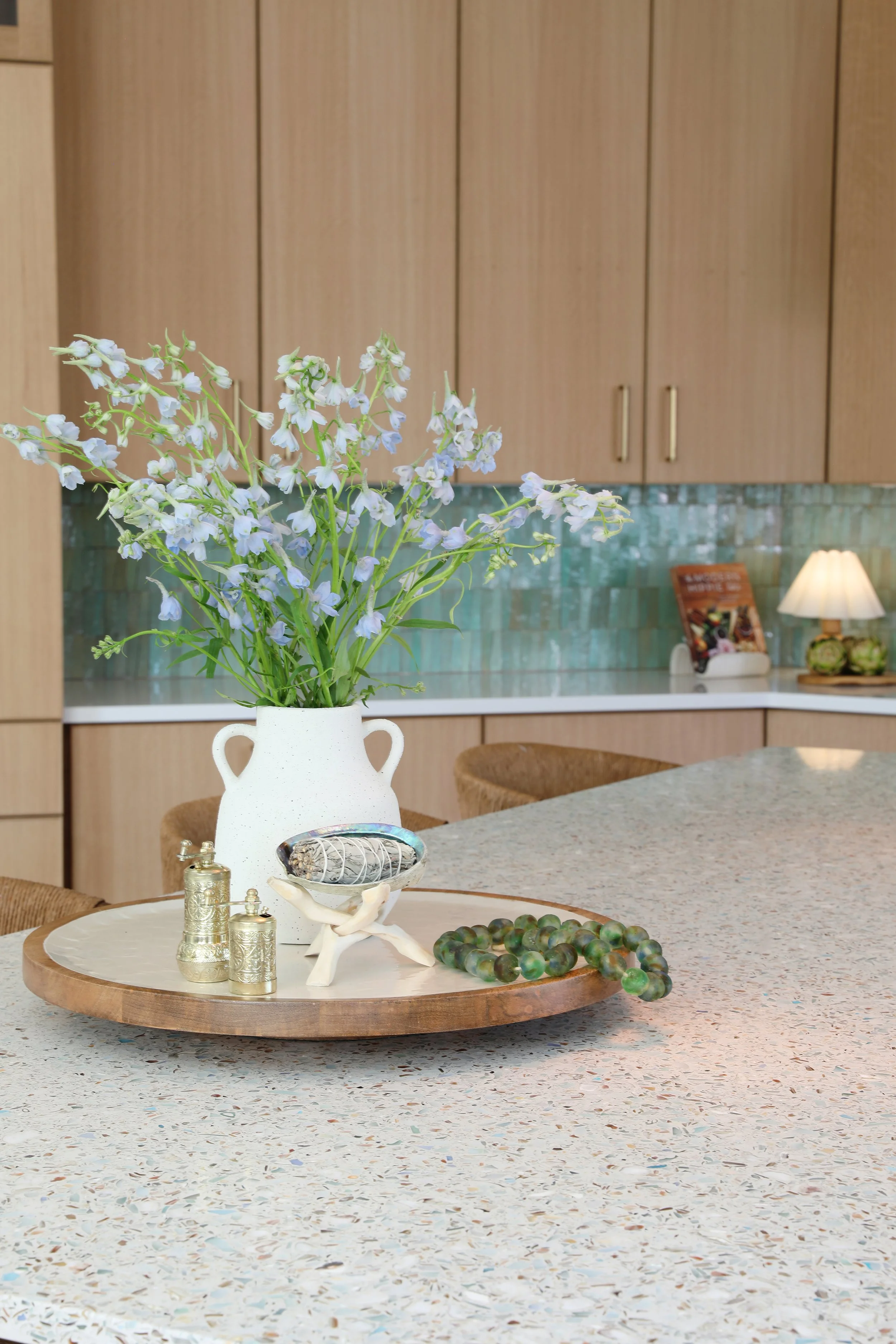 A kitchen with wood cabinets and a tile backsplash, with a countertop and several objects including a white pitcher with blue flowers, small brass containers, decorative stones, and a beaded necklace on a round wooden tray.