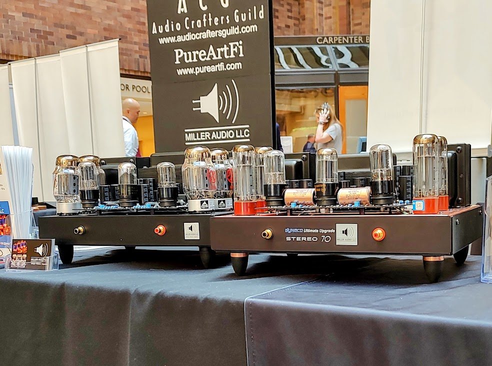 A vintage-style radio or audio amplifier with vacuum tubes displayed on a black cloth-covered table at an exhibition. In the background, there are banners for audio equipment companies and people browsing.
