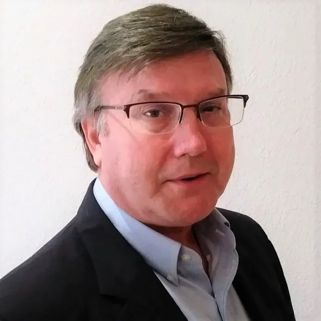 A middle-aged man with glasses, wearing a dark blazer and light shirt, standing against a plain white wall.