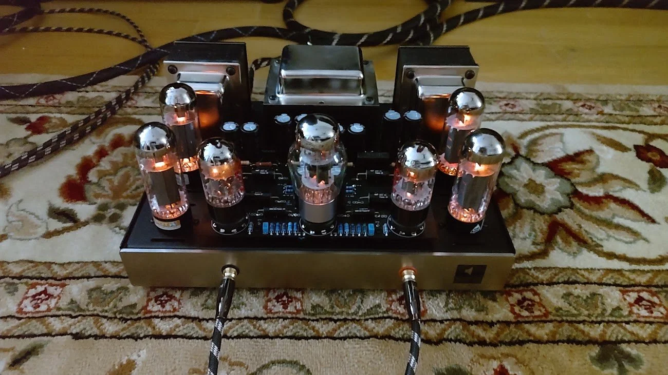A vintage-style tube amplifier with glowing vacuum tubes, placed on a patterned rug with electrical cords in the background.