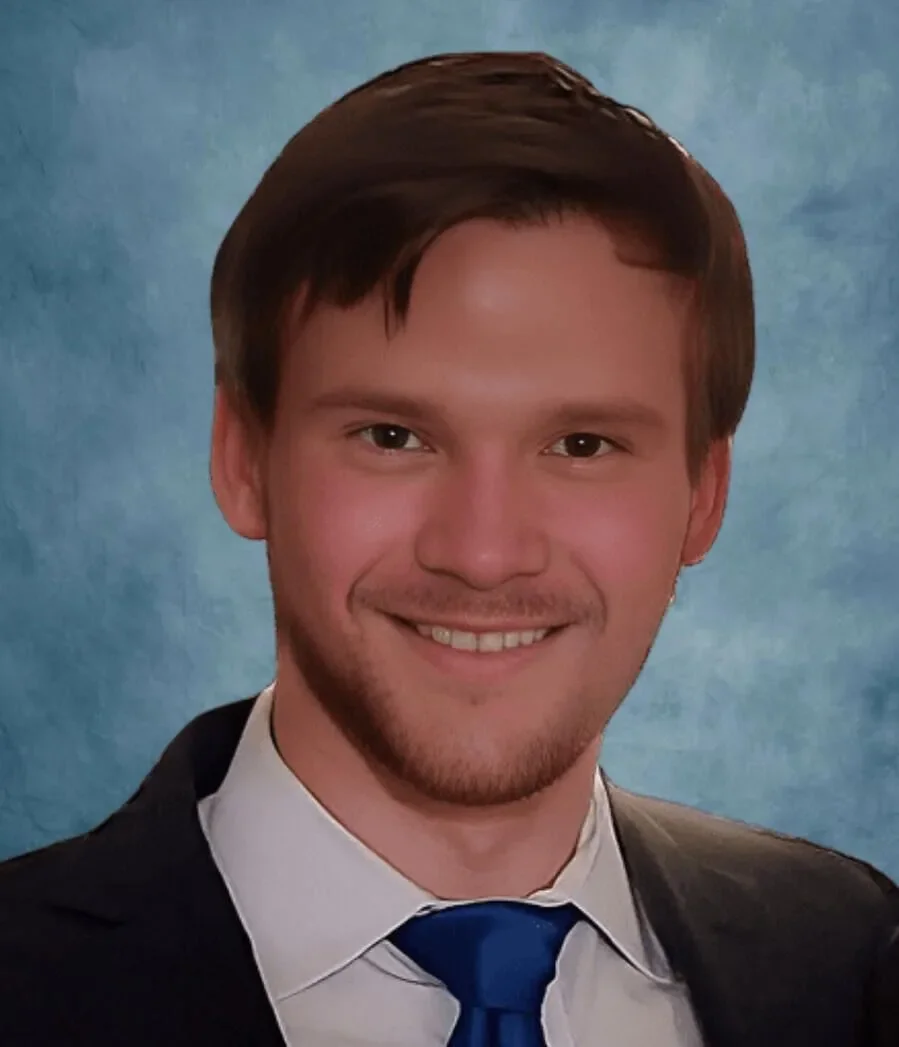 Headshot of a young man with brown hair, smiling, wearing a suit, white shirt, and blue tie against a blue background.