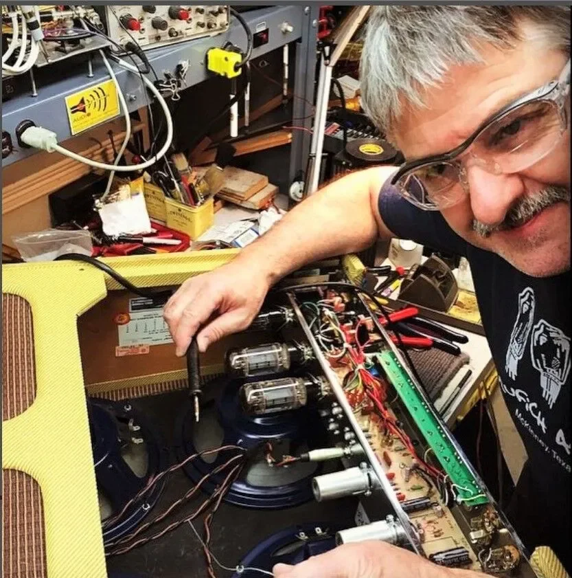 A man with glasses working on the internal electronics of a guitar amplifier in a workshop.