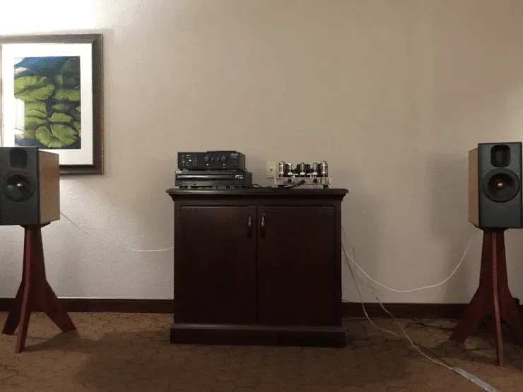 A vintage stereo system with speakers on stands, placed on a dark wooden cabinet in a room with beige walls, a framed picture on the wall, and carpeted floor.