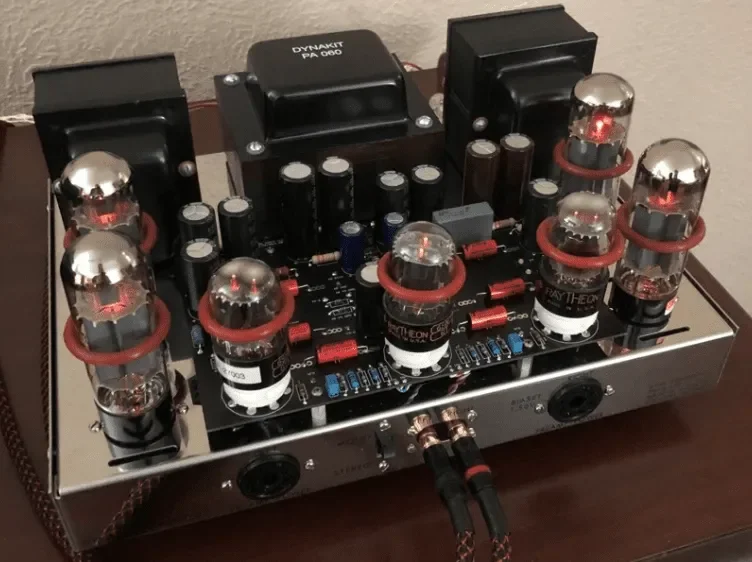 Close-up of a vintage tube amplifier with glowing vacuum tubes, black transformers, and a metallic chassis on a wooden surface.