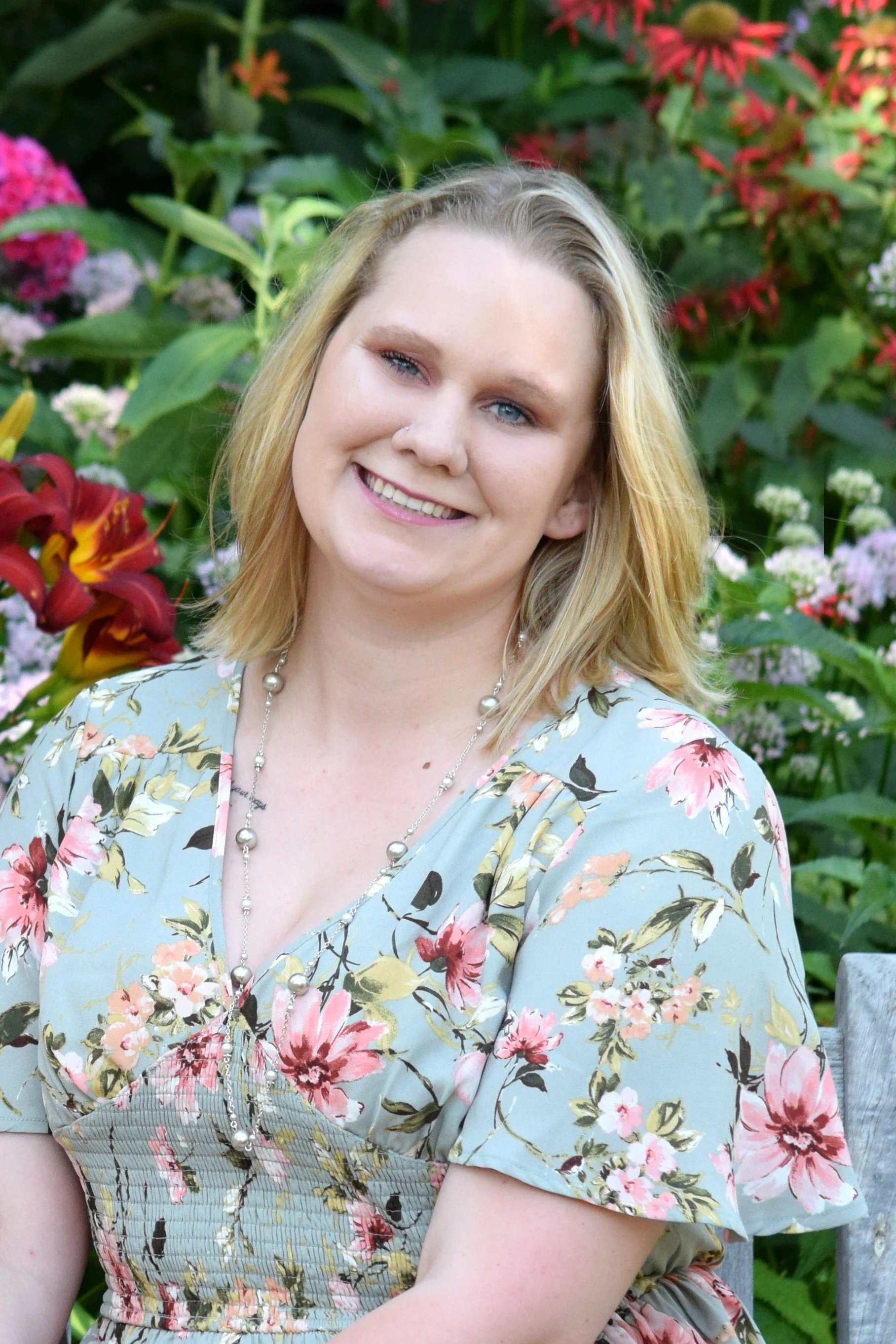 A woman with blonde hair smiling in front of a colorful garden with pink, red, and white flowers.