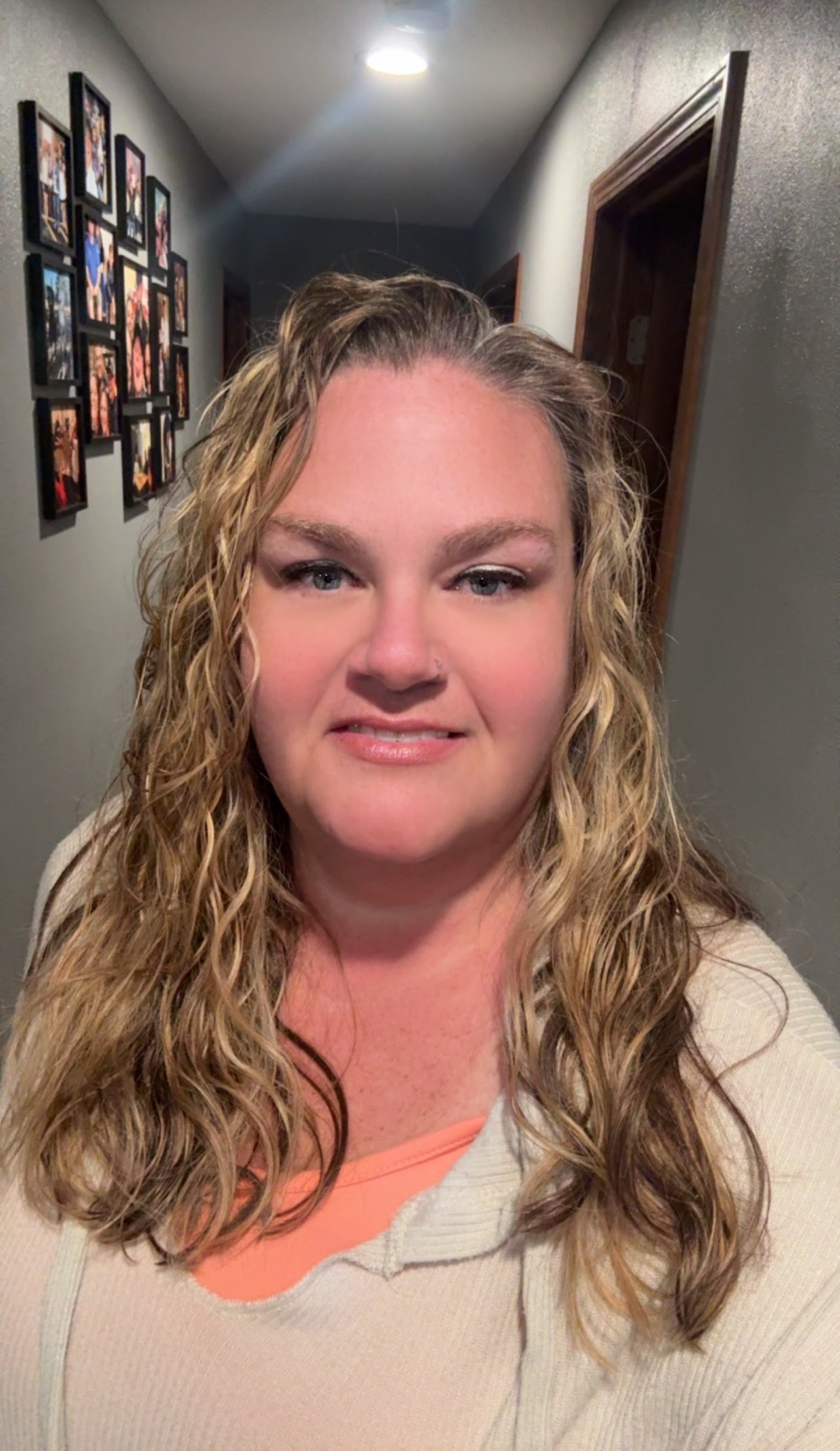 A woman with long, curly blonde hair takes a selfie in a hallway with framed photos on the wall.