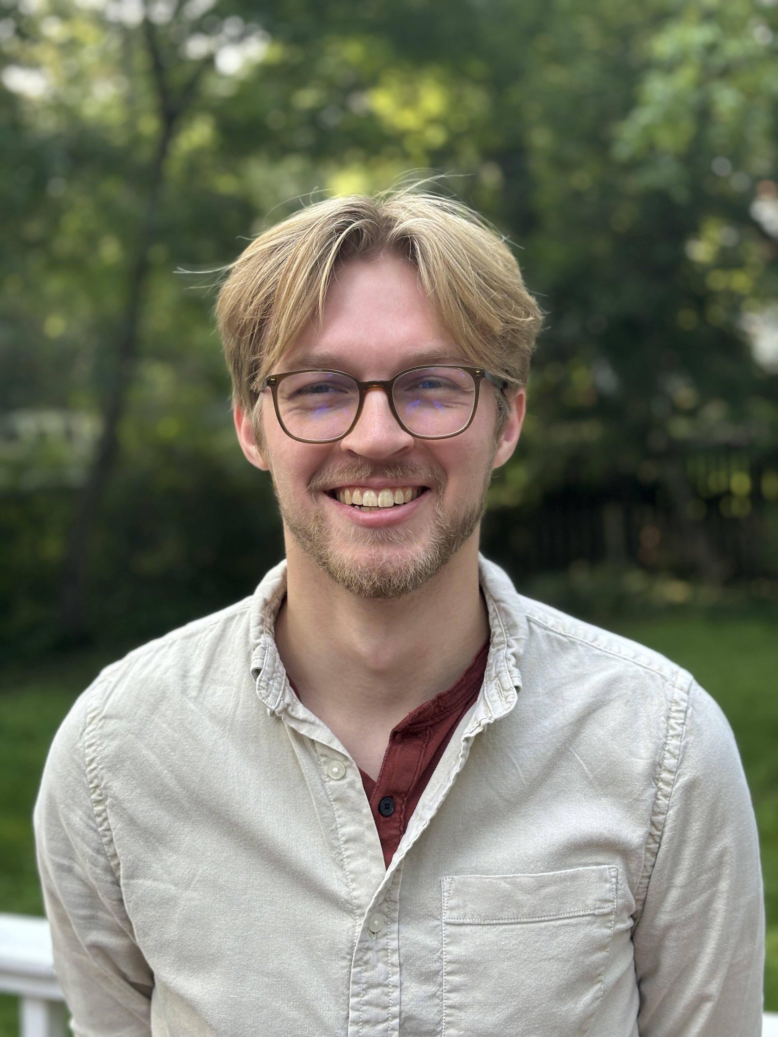 Portrait of a young man with glasses, light brown hair, and a beard, smiling outdoors in front of green trees.