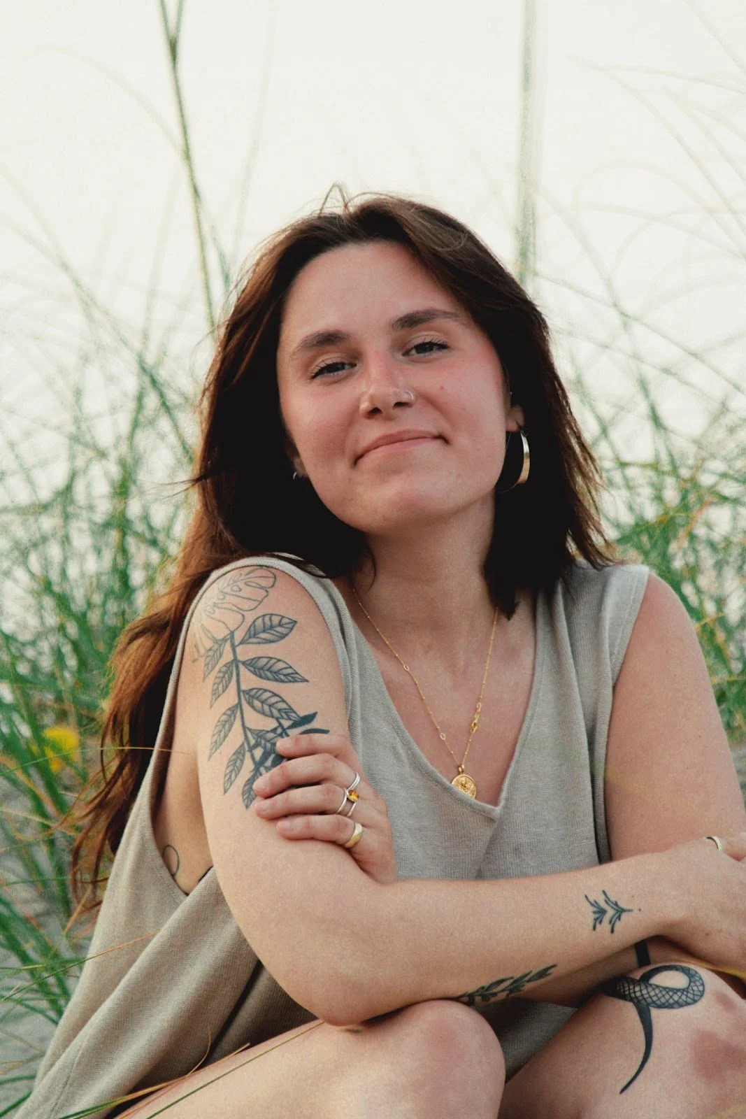 A woman with shoulder-length dark brown hair and tattoos on her arms, sitting outdoors among tall grasses, wearing a sleeveless beige top, jewelry, and earrings.