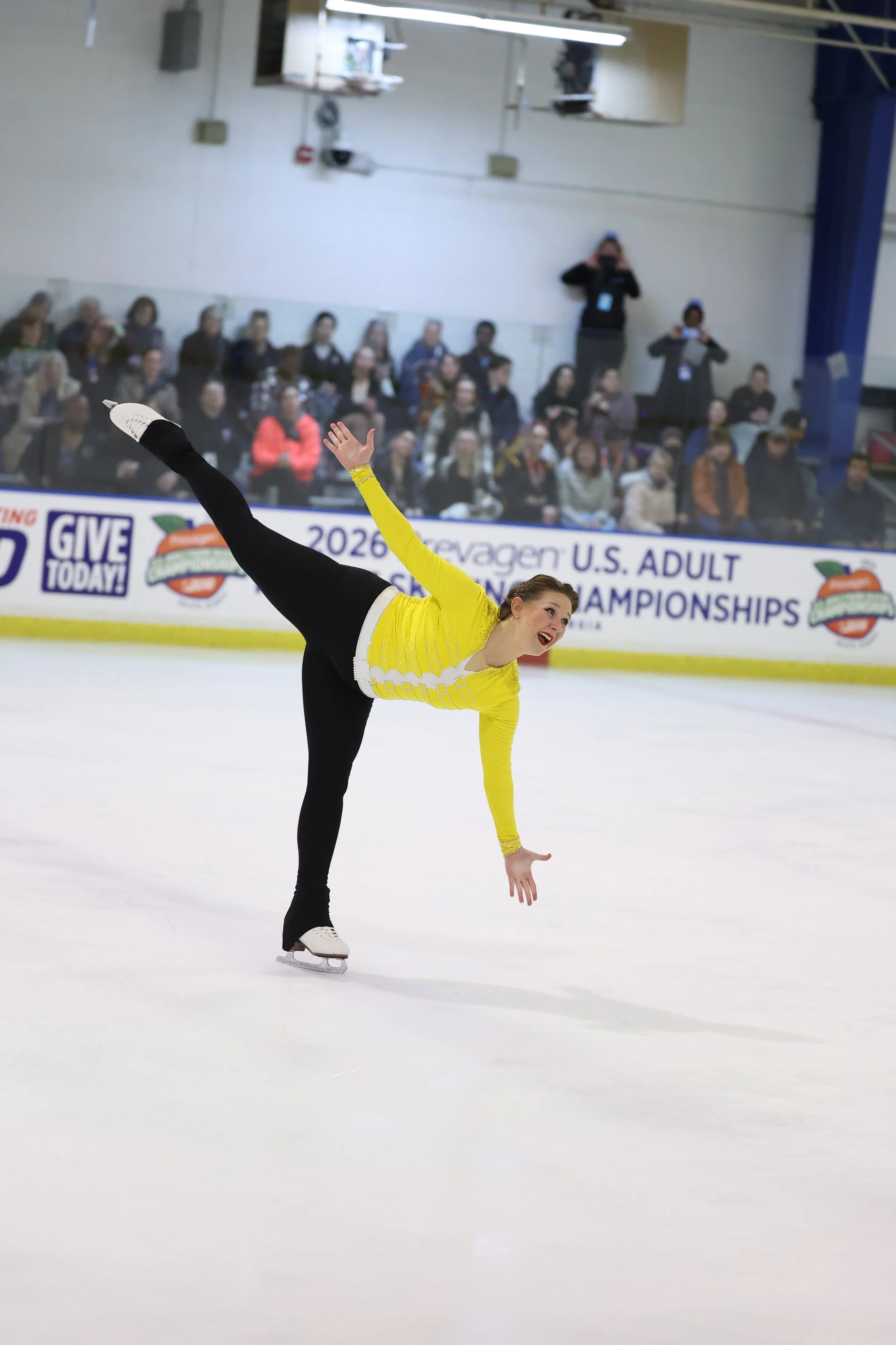 Female figure skater in a yellow top and black pants performing on ice rink during a competition, with an audience in the background and a person taking a photo.
