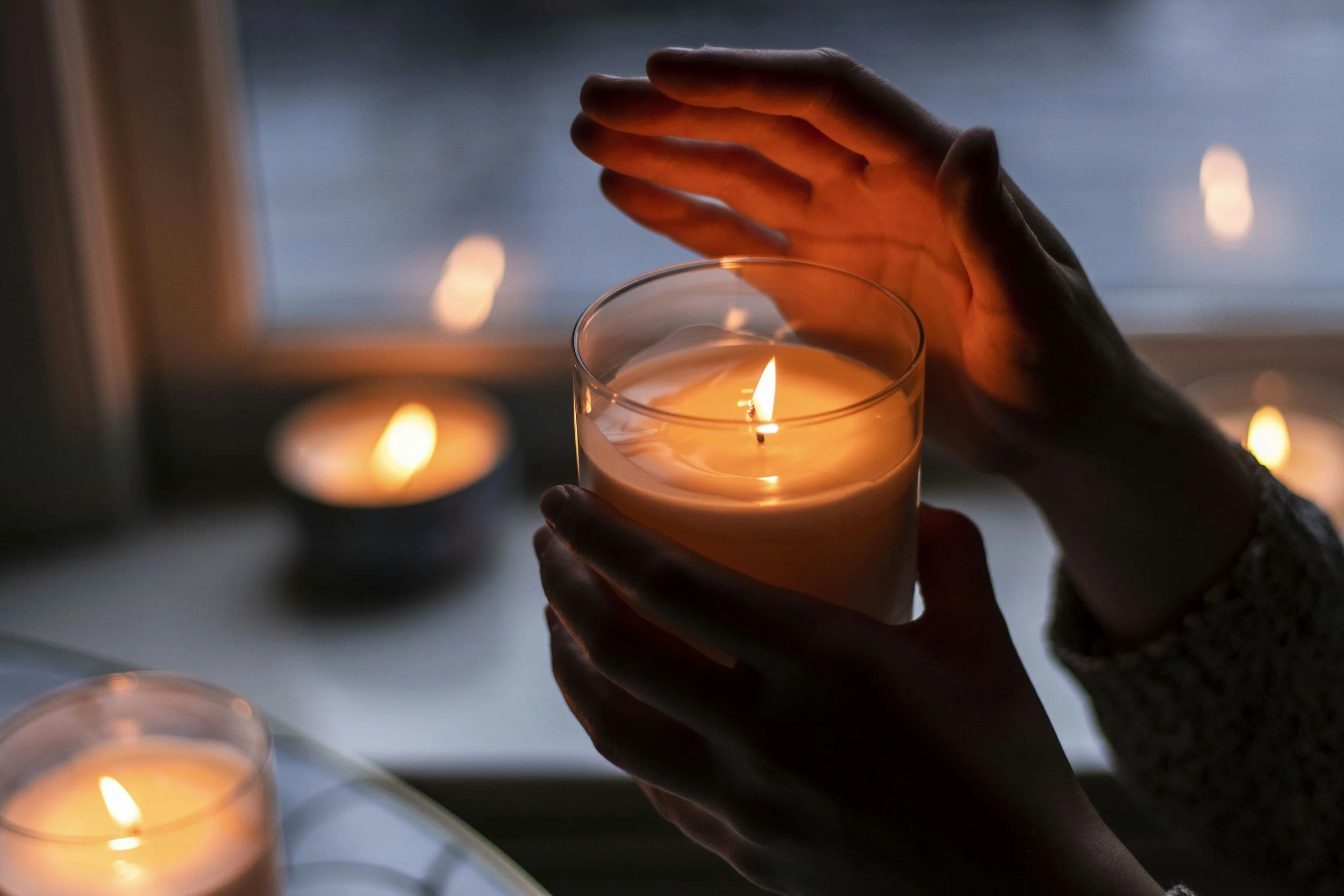 Person holding a lit candle in a glass jar during dusk or evening with other blurred candles in the background.