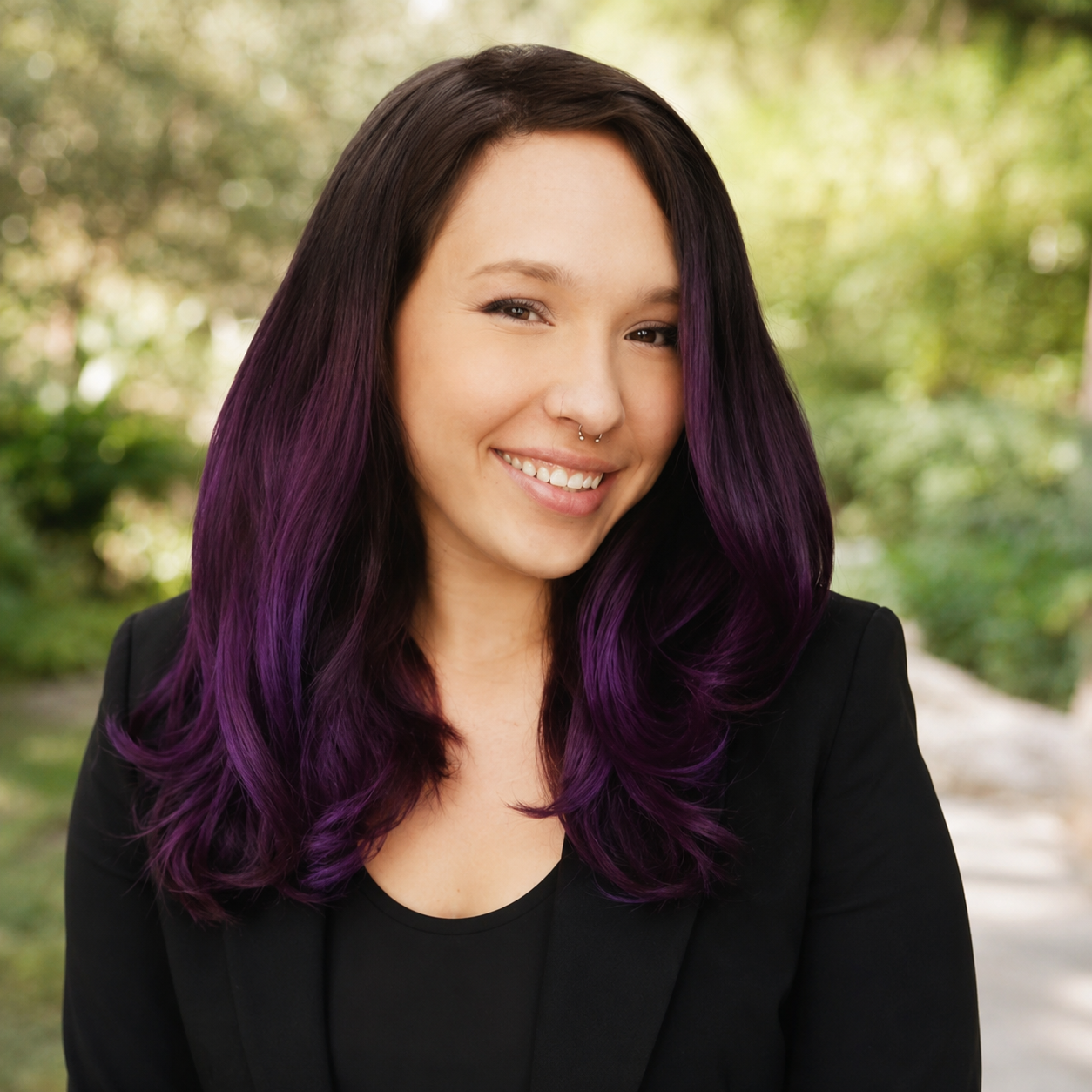 A young woman with shoulder-length black hair with purple highlights, smiling outdoors with a blurred green background, wearing a black blazer and top.