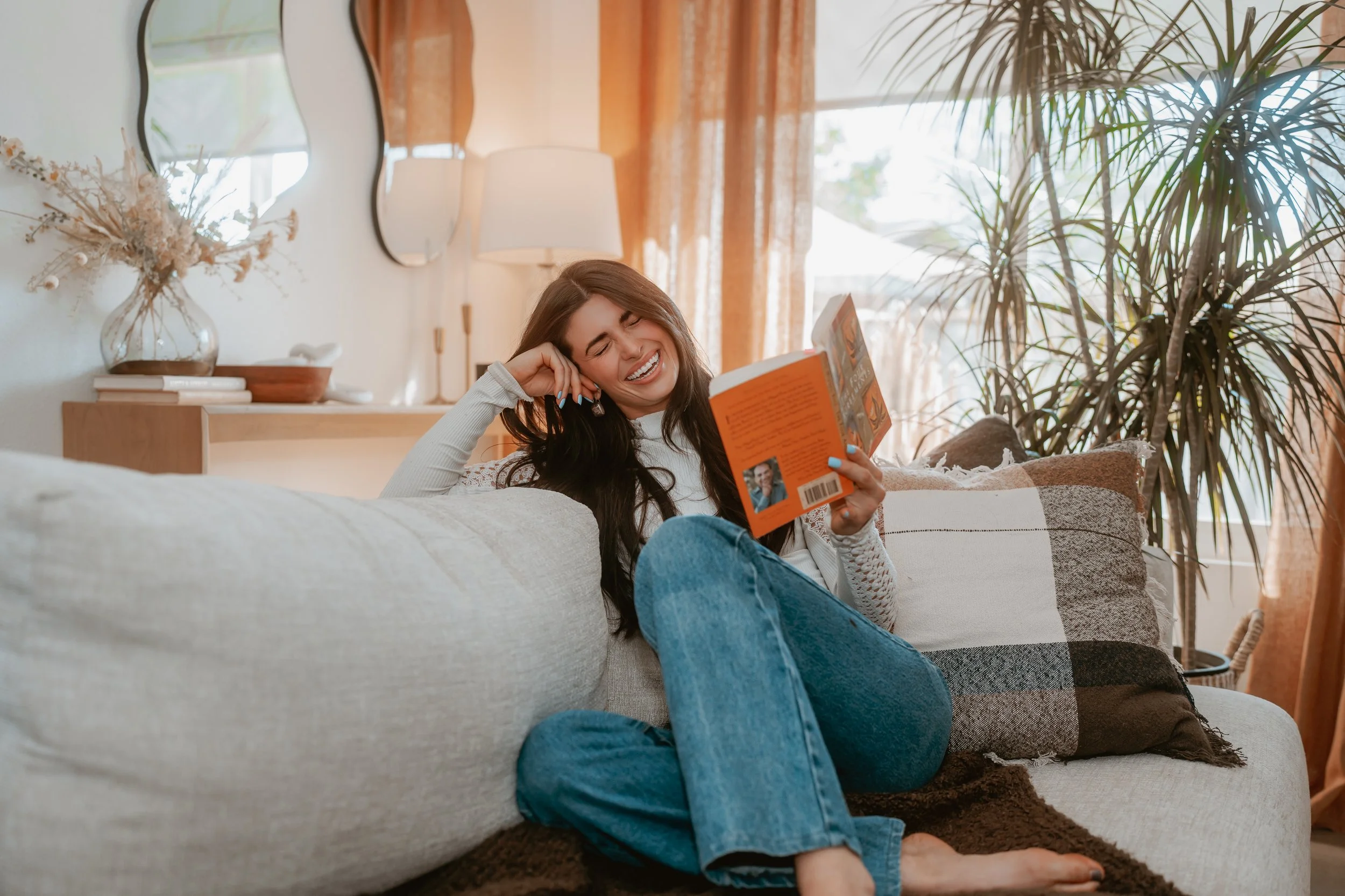 A woman with long dark hair is sitting on a couch, smiling and reading a book, with her eyes closed. The room is bright and cozy, decorated with a large plant, a mirror, and a lamp, with sunlight coming through a window with curtains.