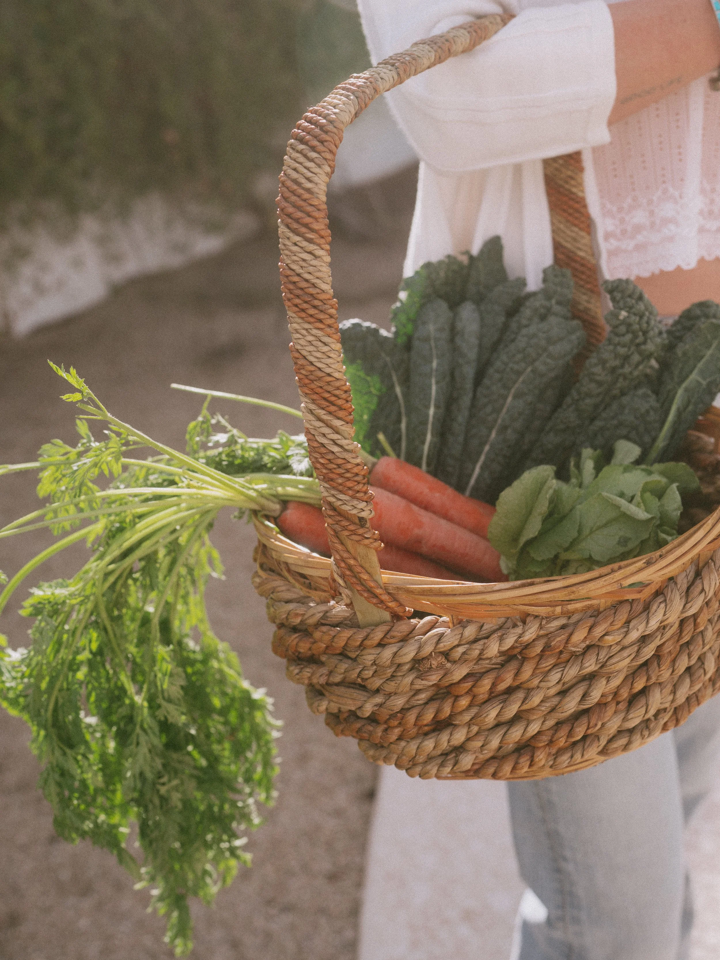 A woven basket filled with fresh vegetables including carrots and leafy greens, set on a surface with a piece of white cloth nearby.