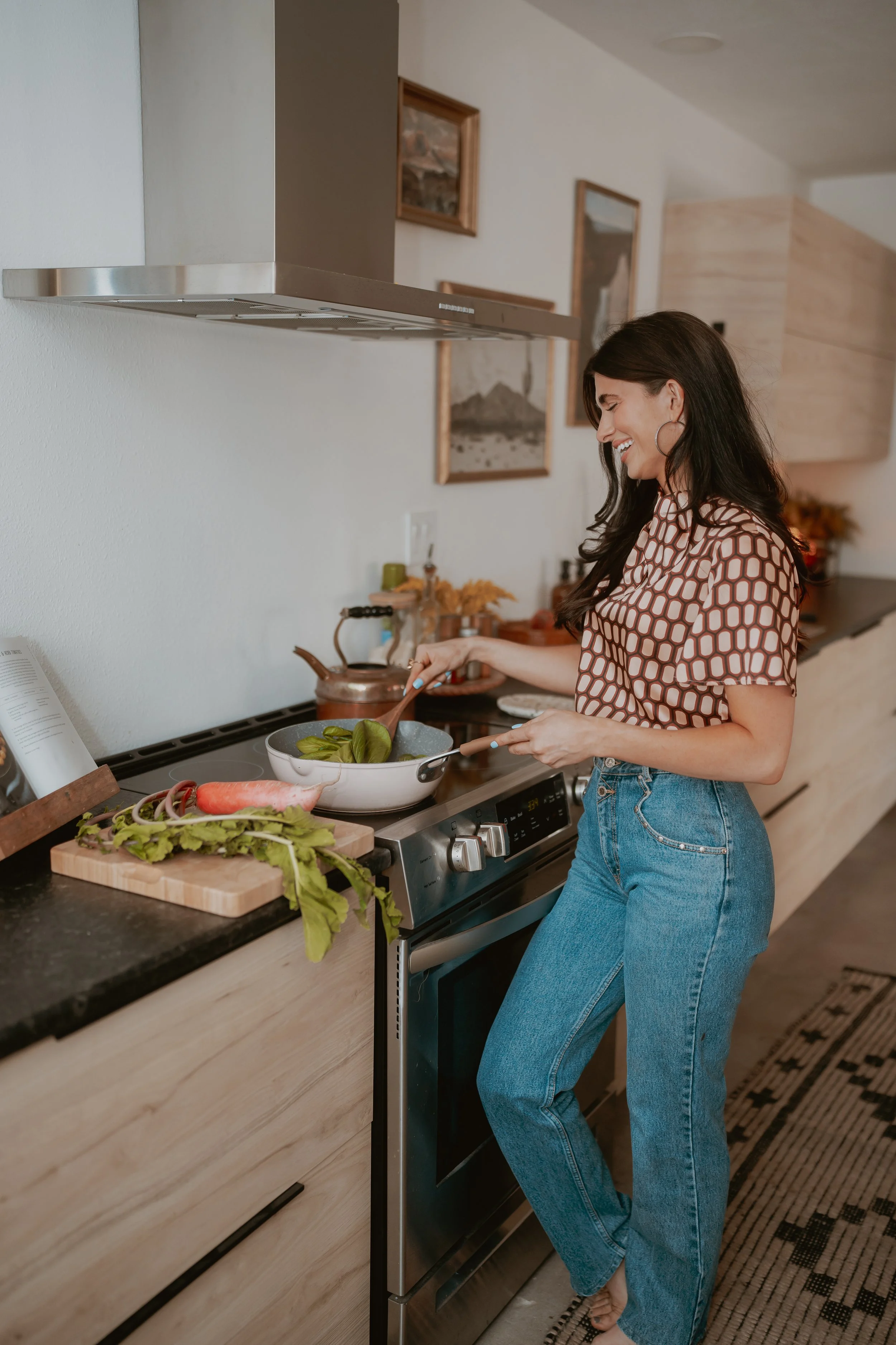 A woman with long dark hair, hoop earrings, a patterned top, and blue jeans is cooking in a modern kitchen. She is standing at a stove, smiling, and stirring a pot with vegetables. The kitchen has a wooden cabinet, colorful artwork on the wall, and various cooking utensils and ingredients on the counter.