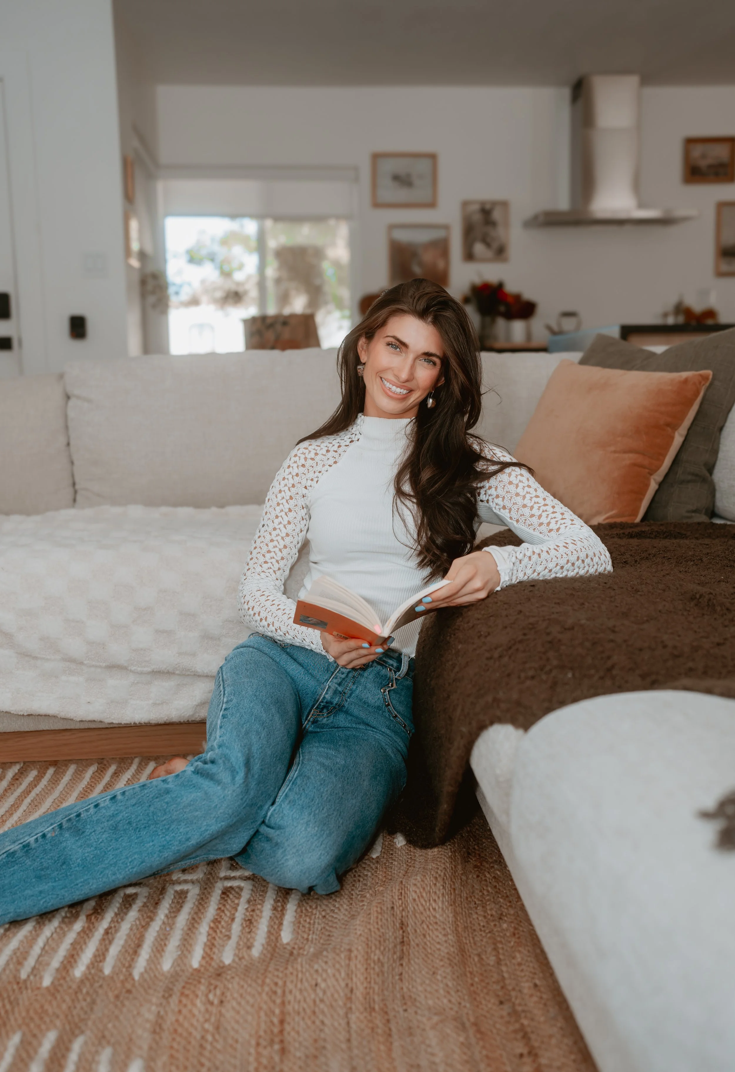 A woman sitting on the floor leaning against a sofa, smiling and holding a book in a living room with paintings on the wall and a large window in the background.