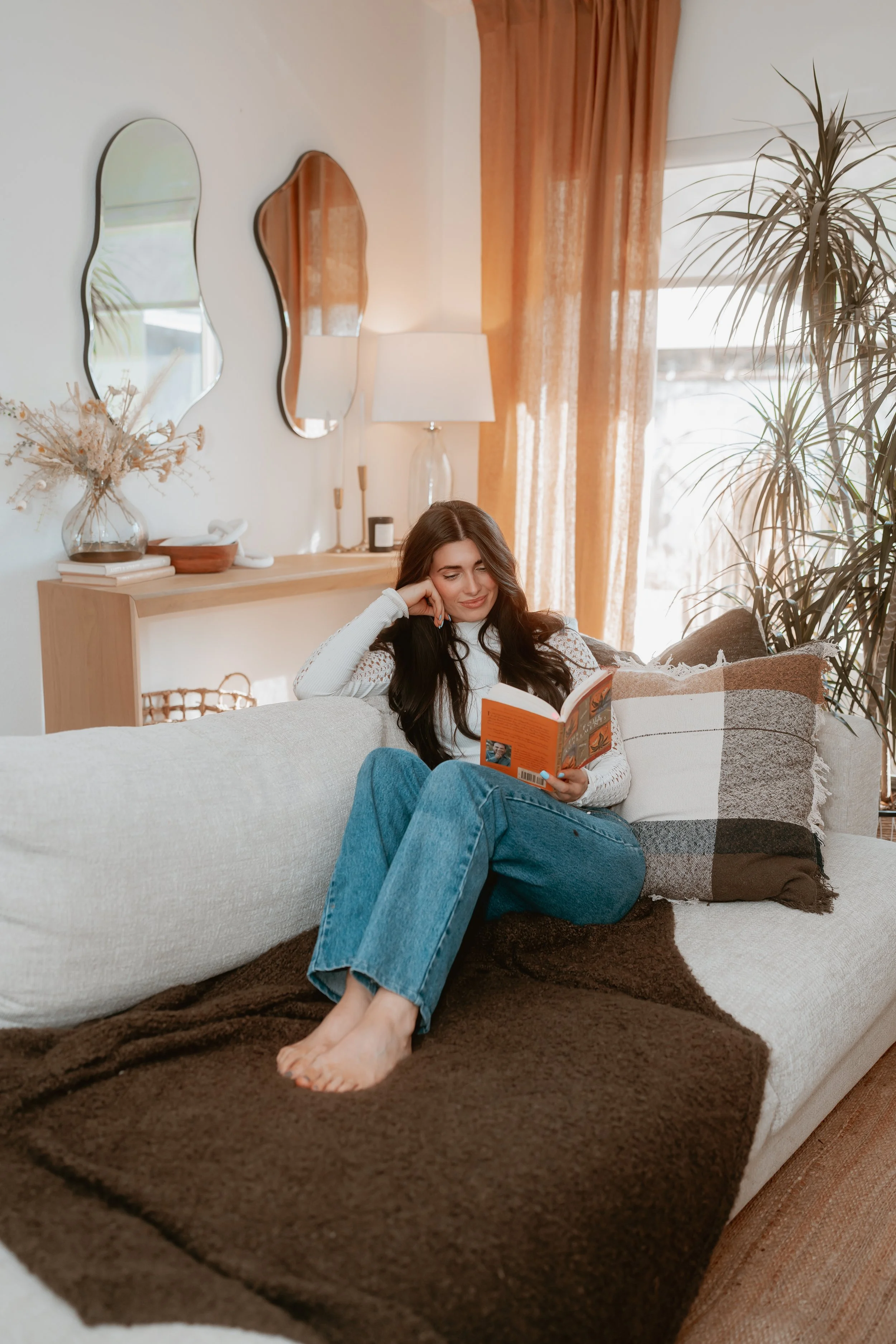 A young woman with long dark hair is relaxing on a beige couch, reading a book. She is barefoot, wearing blue jeans and a white long-sleeve top. The living room has warm lighting, with orange curtains, a large potted plant, and decorative mirrors on the wall.