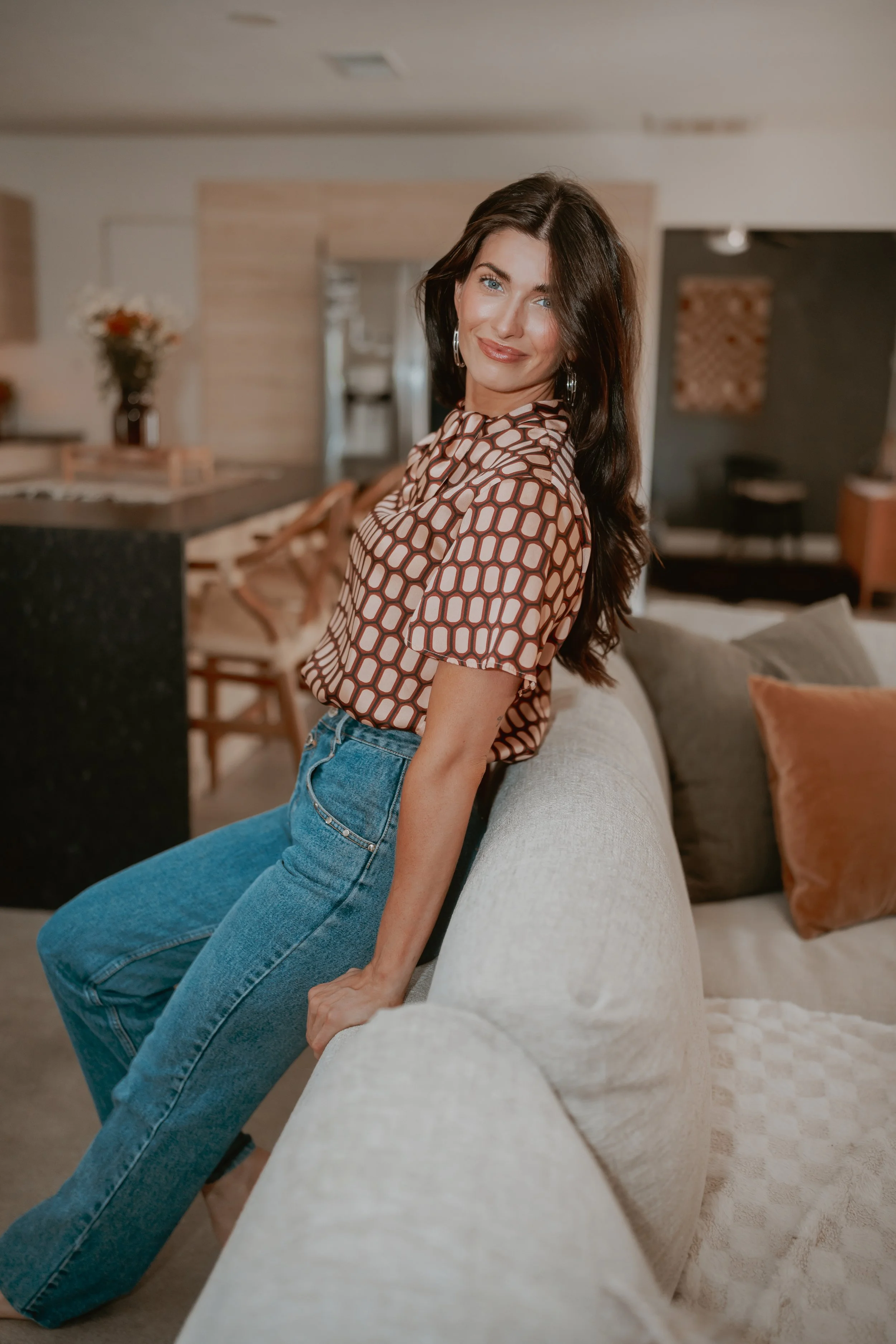 A woman with long dark hair and blue eyes sitting on a cream-colored sofa in a cozy, well-decorated living room, smiling at the camera.