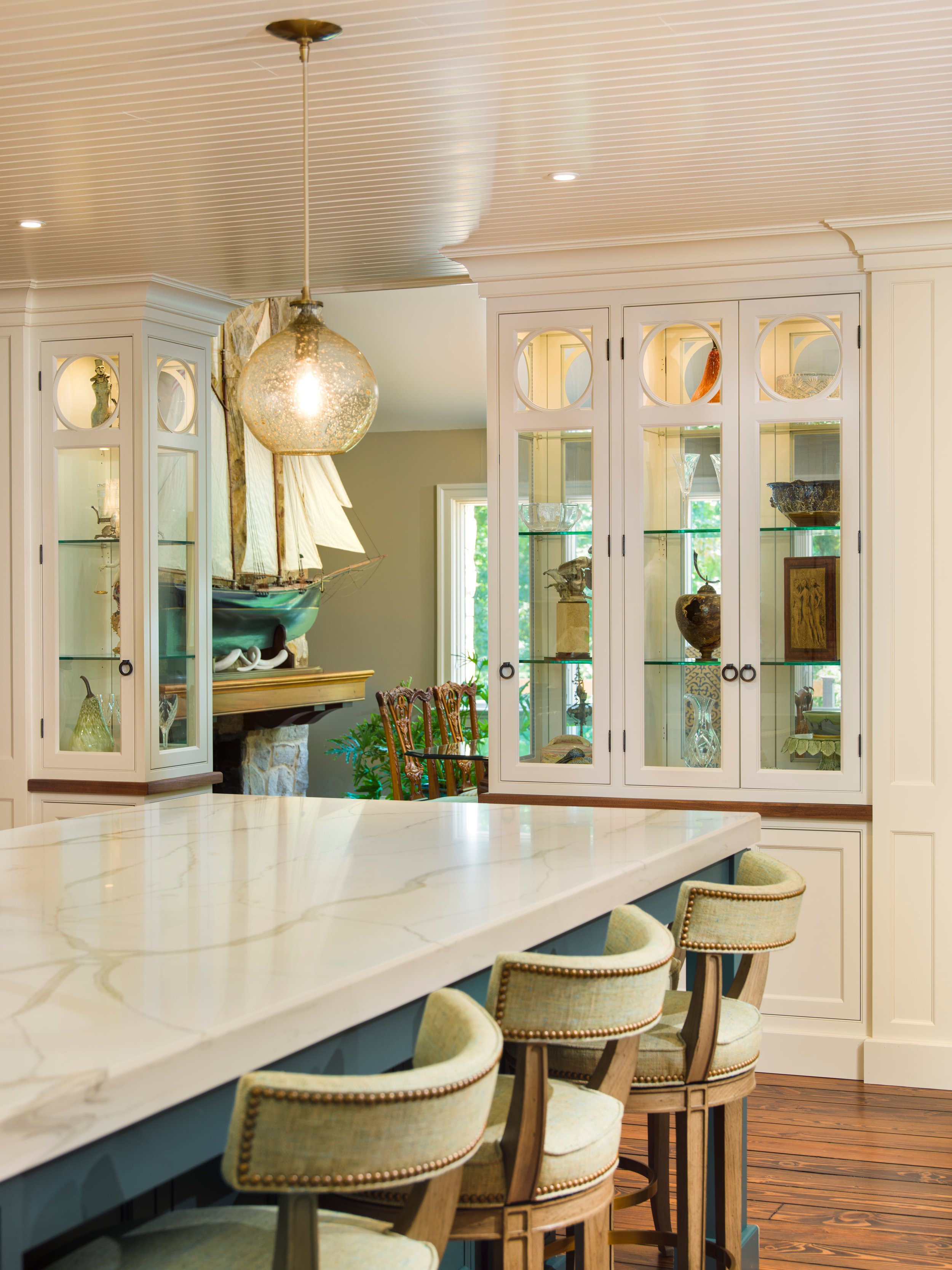 Kitchen island with cream-colored marble countertop and beige upholstered chairs, white cabinets with glass doors displaying decorative items, hardwood floors, large window, cream walls, with a lit globe pendant light hanging from the ceiling.