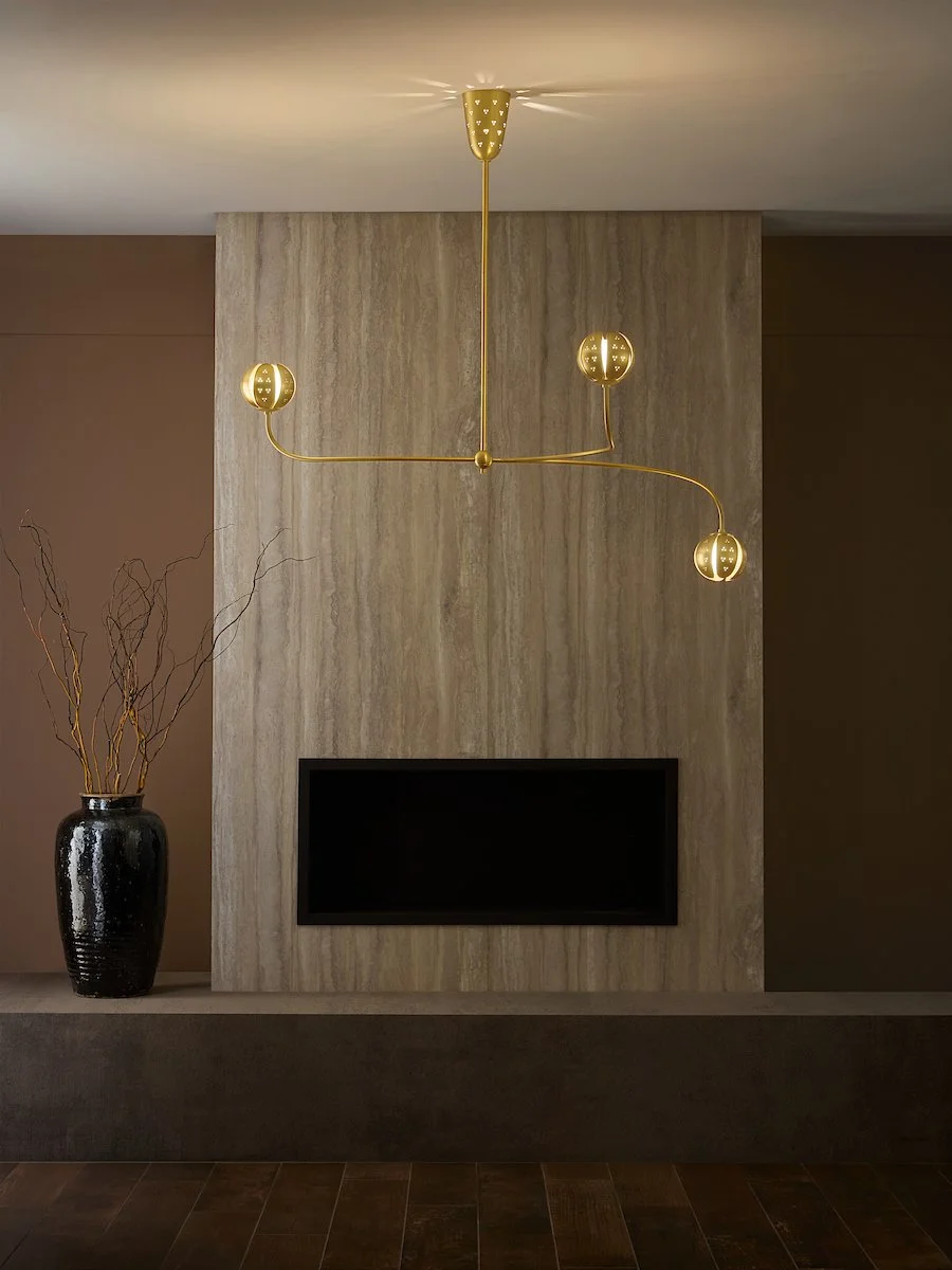 Modern living room with a gold chandelier hanging over a beige stone accent wall, a black flat-screen TV inset into the wall, and a black vase with dried branches on a ledge.