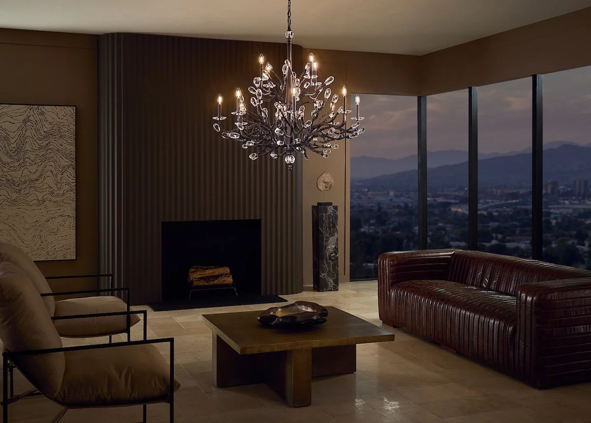 Living room with a large chandelier, tan armchairs, a dark leather sofa, a wooden coffee table, a modern fireplace, and cityscape view through floor-to-ceiling windows at dusk.