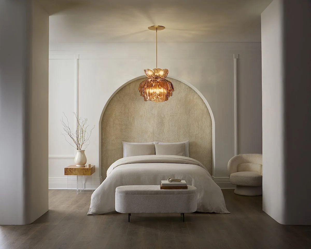 Minimalist bedroom with a white bed, beige fabric headboard, and a small bench across the foot of the bed. Decor includes a white vase with twigs on a floating wooden shelf, a round beige chair, and a pendant light with a warm tone hanging from the c