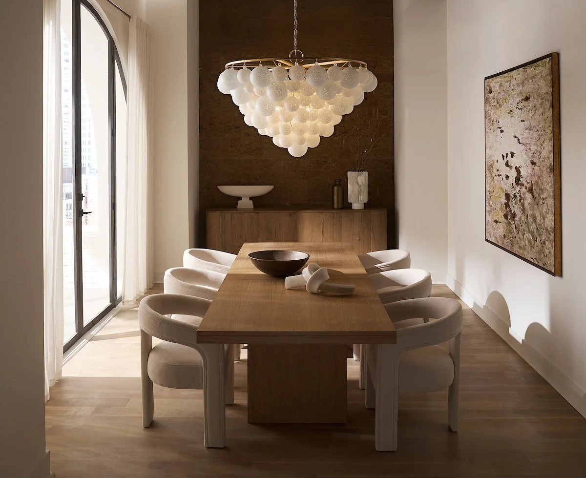 Modern dining room with a wooden table, six white chairs, a chandelier with white globes, and a sliding glass door with curtains. Artwork on the wall and decorative vases on a sideboard.