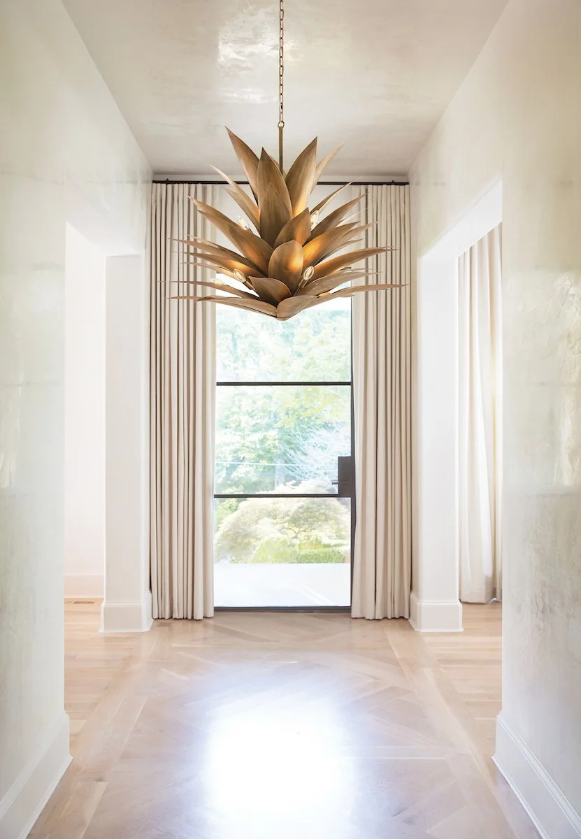 A modern chandelier designed like overlapping leaves hanging in a bright hallway with light-colored walls, curtains, and hardwood flooring.