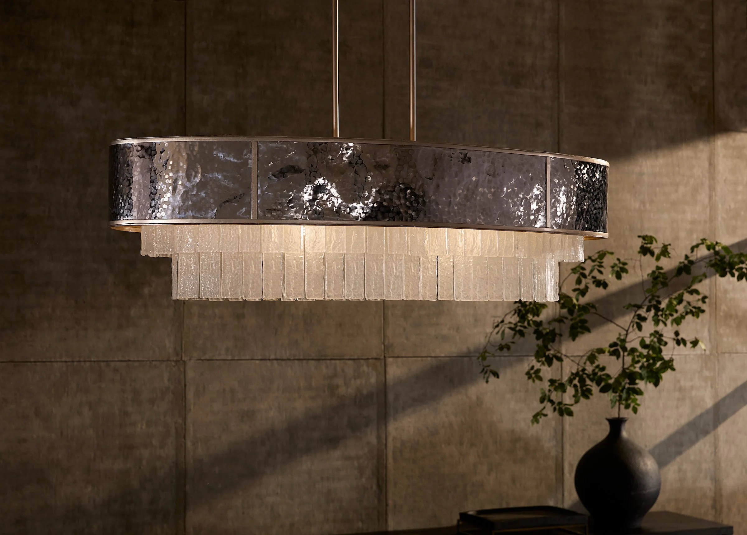 A modern chandelier with a circular band of reflective, textured metal and a frilled glass or acrylic shade, hanging inside a room with wood-paneled walls and a potted plant nearby.
