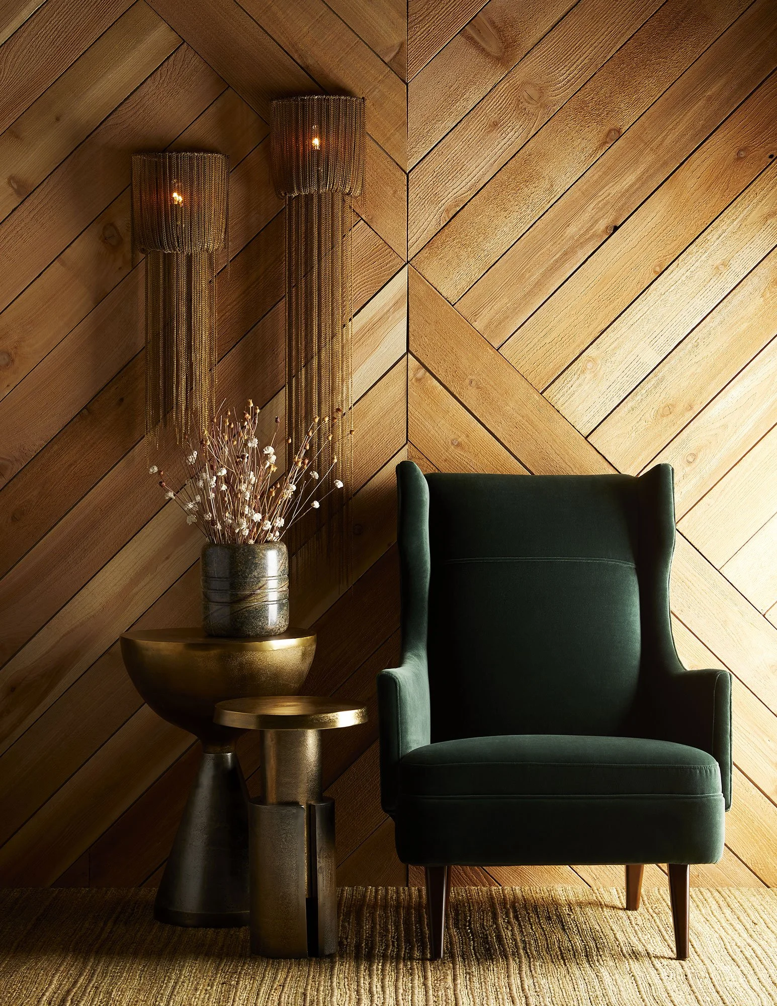 A green upholstered armchair next to a gold-colored side table with a metallic vase of dried white flowers. Two long brass chain-link pendant lights are hanging on a wooden wall with diagonal planks.