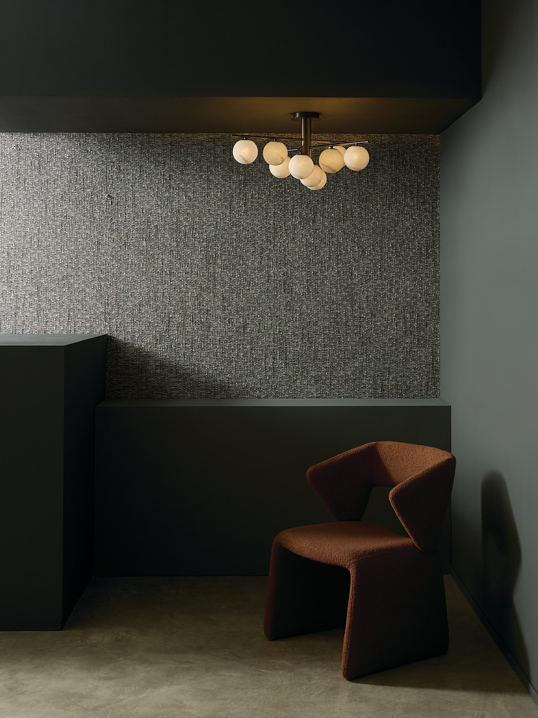 A corner of a room with a textured gray wall, a black ceiling, a modern chandelier with white globe lights, and a striped brown armchair.