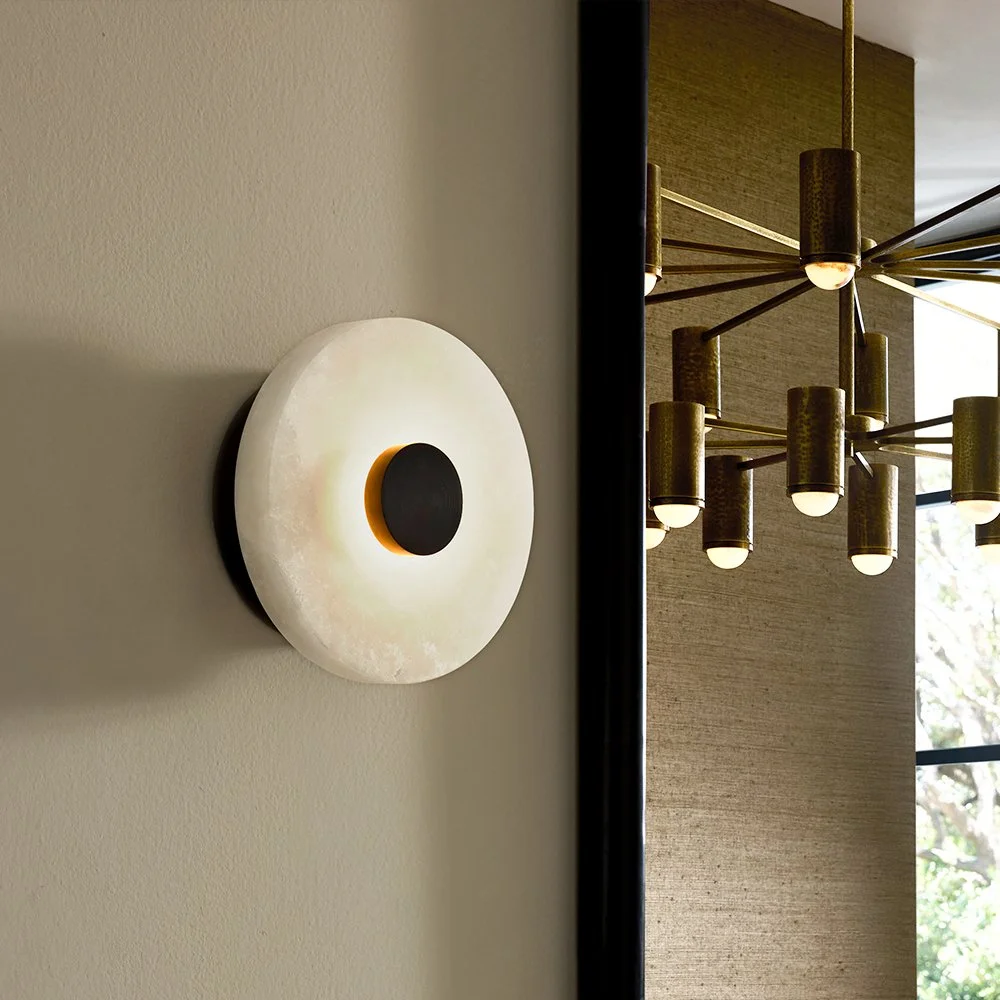 Modern wall sconce light fixture with a circular white shade and a black circular accent, placed on a beige wall; in the background, a contemporary chandelier with multiple gold cylindrical lights hangs from the ceiling near a window.